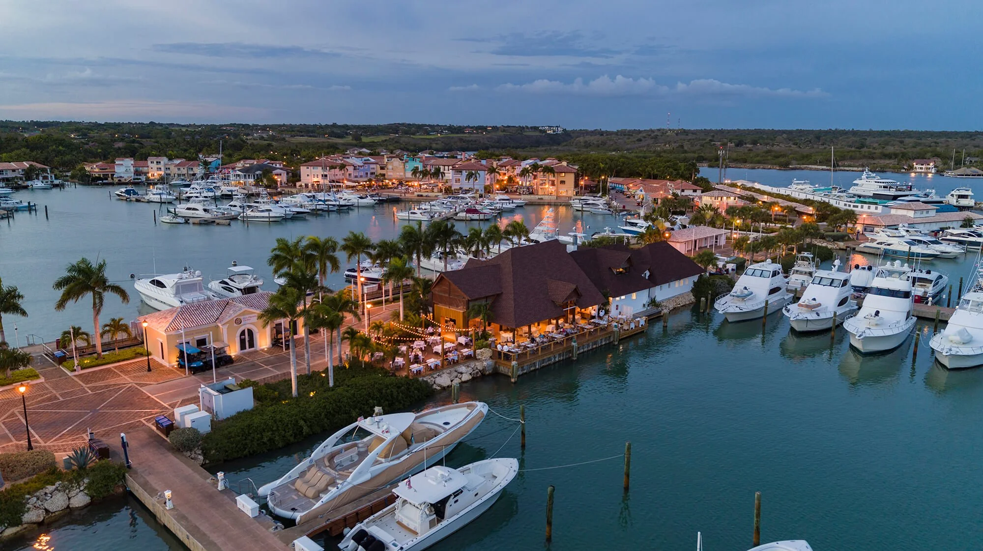 Aerial view of a marina with various yachts and boats docked, with a waterfront restaurant and palm trees illuminated at dusk
