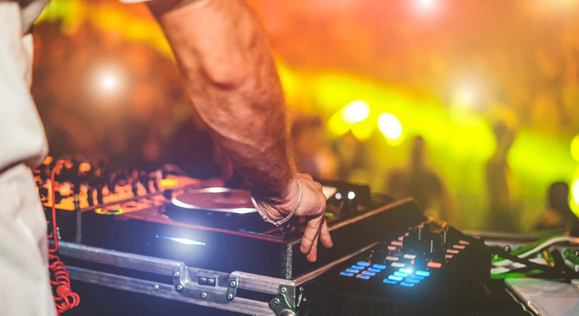 Close-up of a DJ's arm and hand operating a DJ controller at a party or club, with vibrant colorful lights in the background.