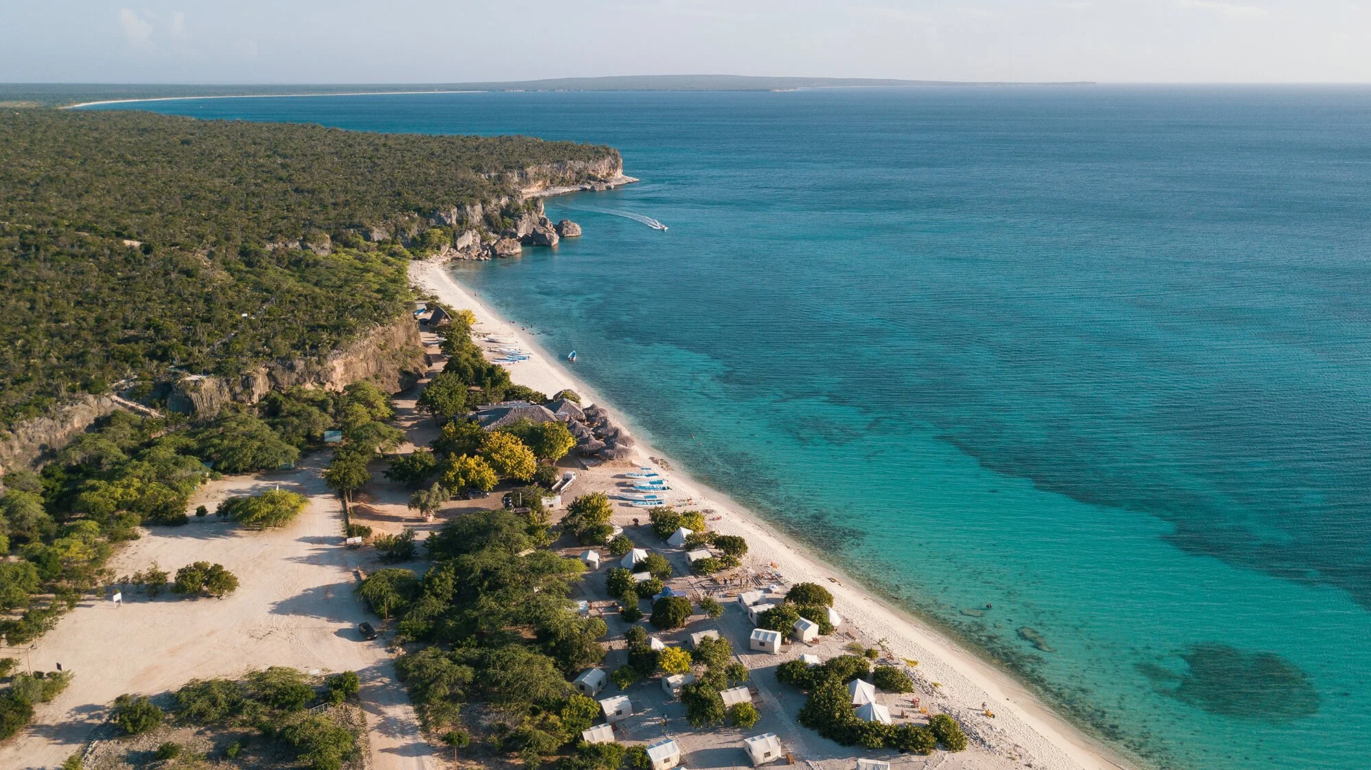 Aerial view of a beachside resort with small tents and trees on the sandy shore, overlooking clear blue ocean water with a distant horizon.