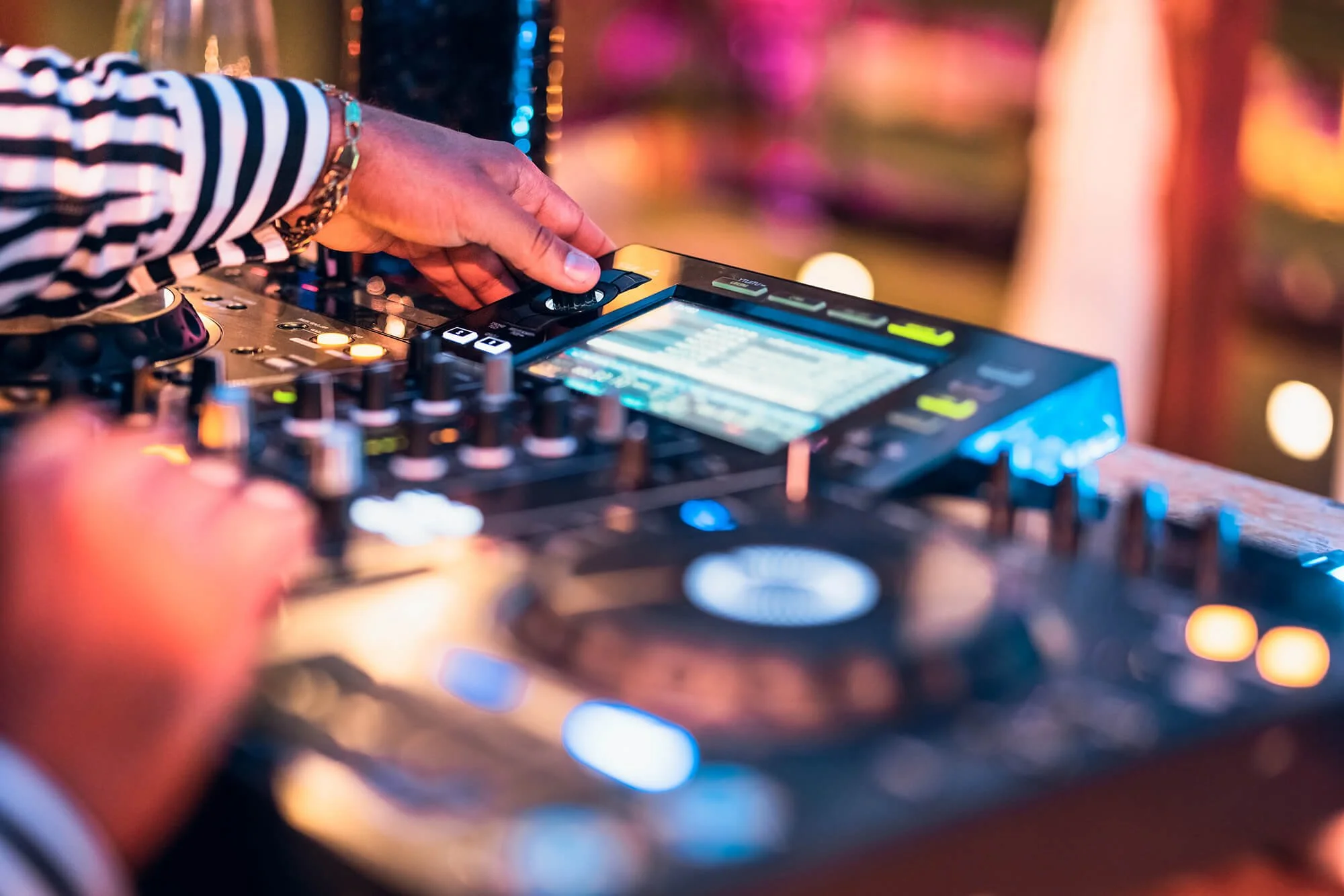 Close-up of a DJ's hands operating a professional DJ controller at a vibrant party or club, with colorful lights in the background.