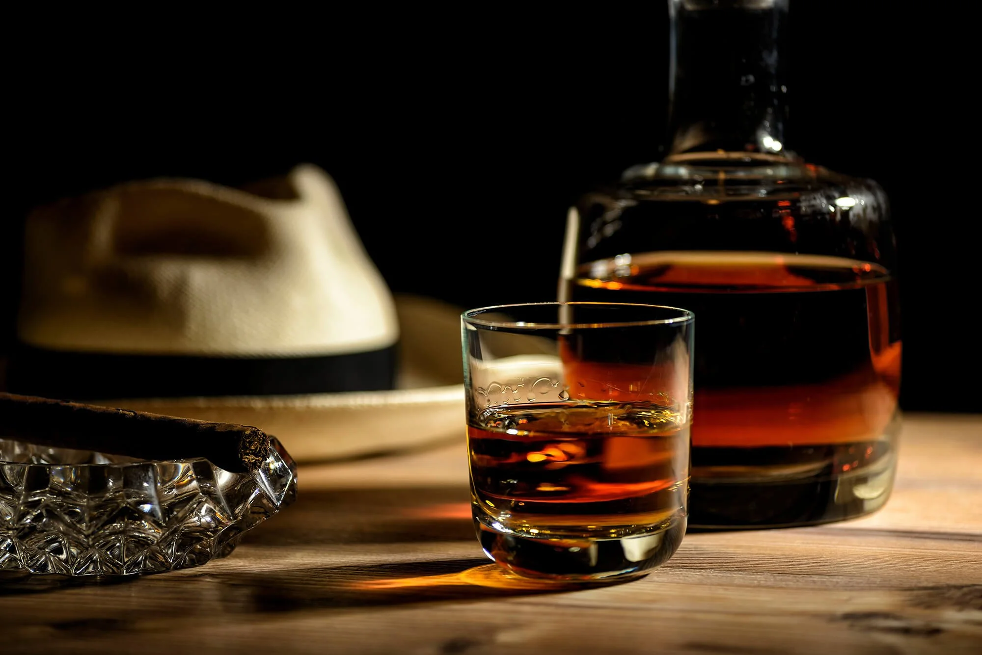 A glass of whiskey being poured from a decanter, with a hat and crystal ashtray on a wooden surface, against a dark background.
