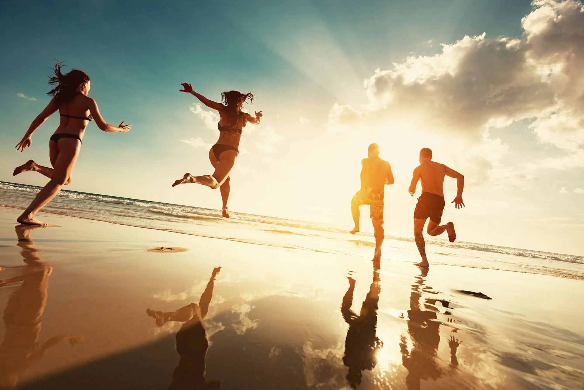 People running and jumping on the beach during sunset, with reflections on the wet sand and a partly cloudy sky.