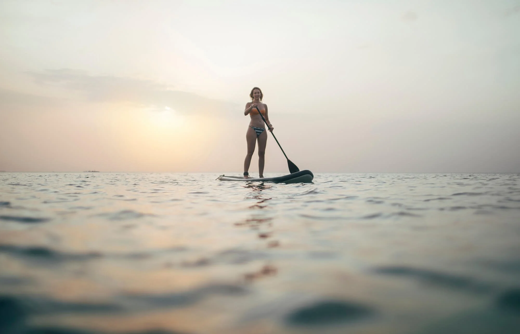 A woman in a bikini stand-up paddleboarding on calm water during sunset or sunrise.