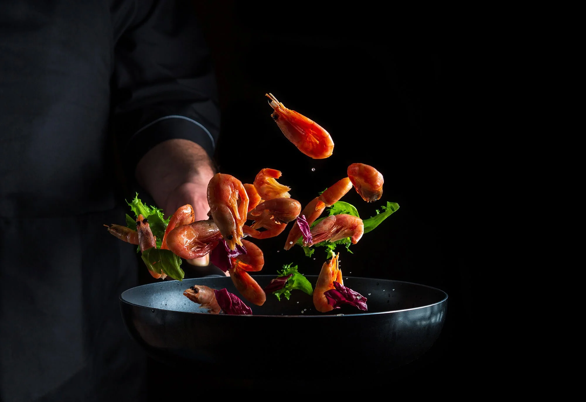 Chef tossing shrimp and mixed greens in a black bowl against a dark background.