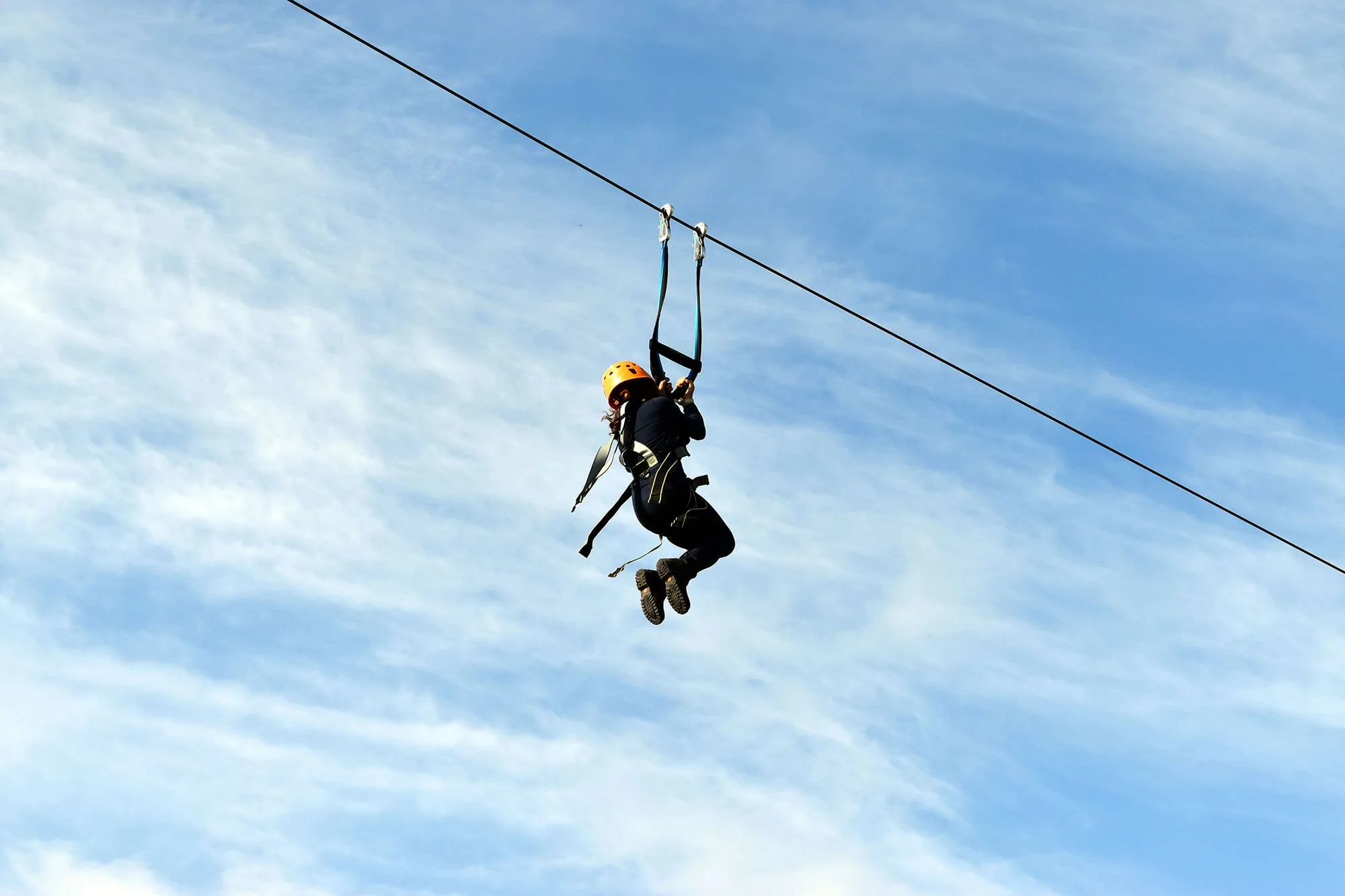 A person wearing a yellow helmet and black safety harness is ziplining through the sky against a background of blue sky and wispy clouds.