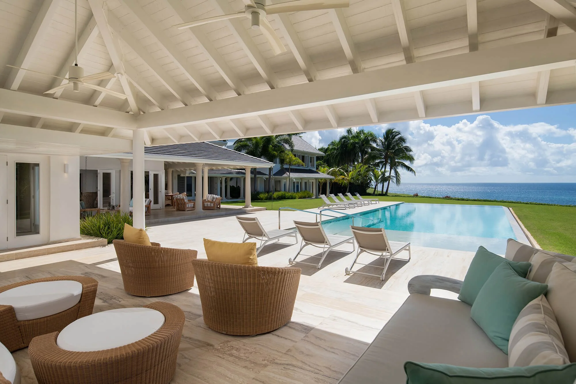 Luxury outdoor pool area with pool chairs, tropical landscaping, and ocean view under a covered patio with white ceiling and fans.