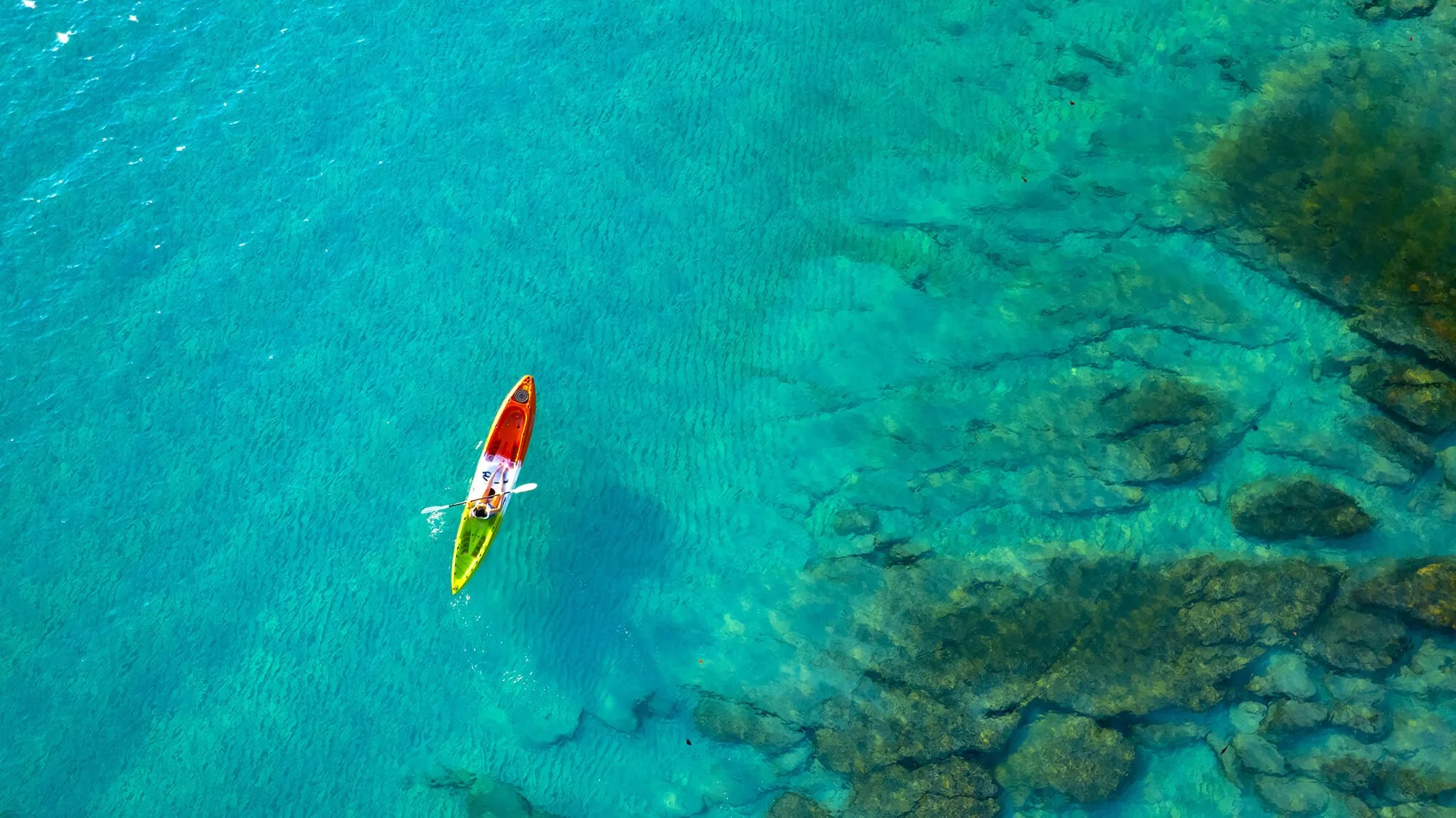 An overhead view of a colorful kayak on clear blue water near rocks.
