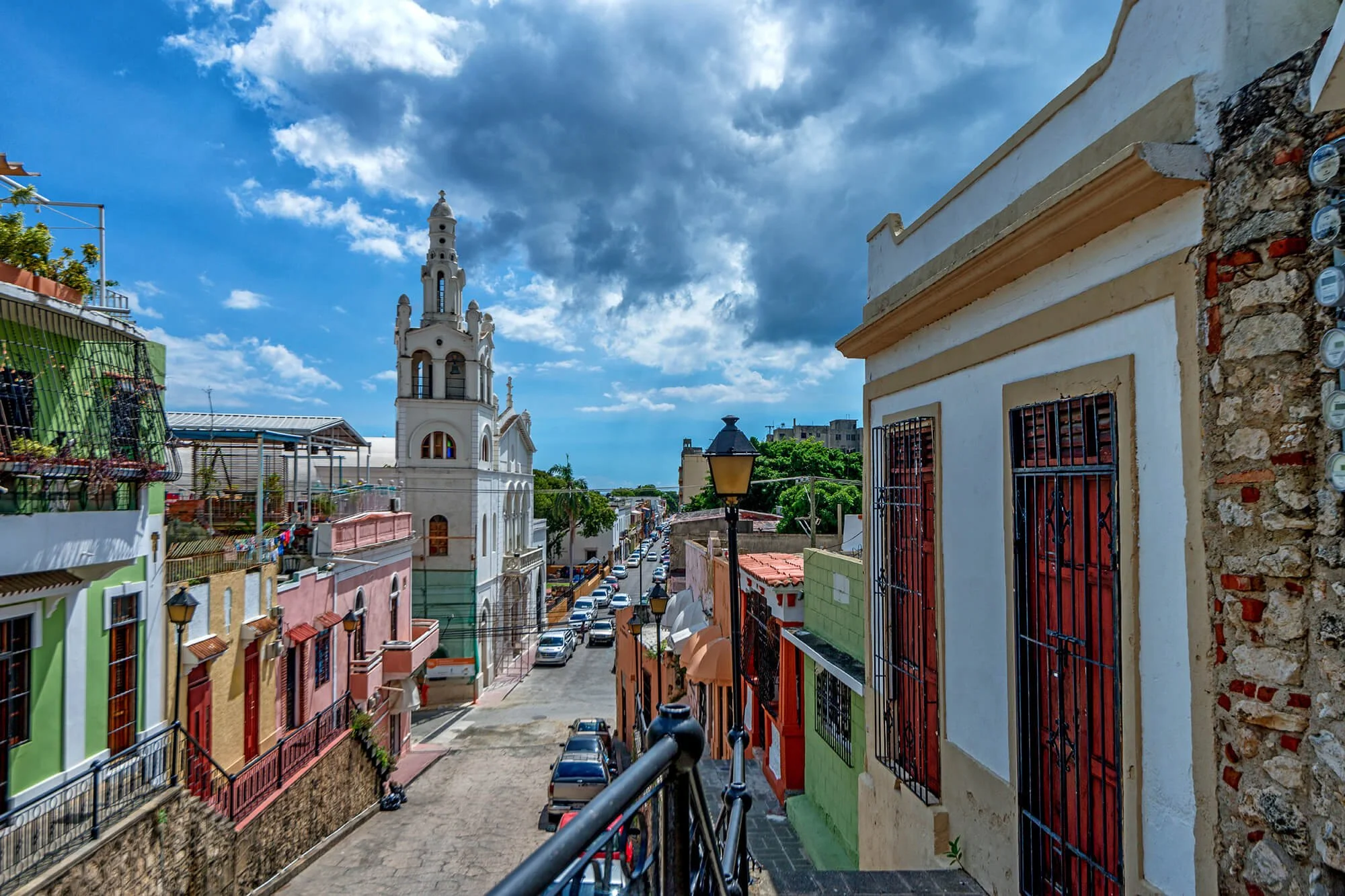 Colorful buildings along a cobblestone street with a white church tower in the background under a partly cloudy sky.