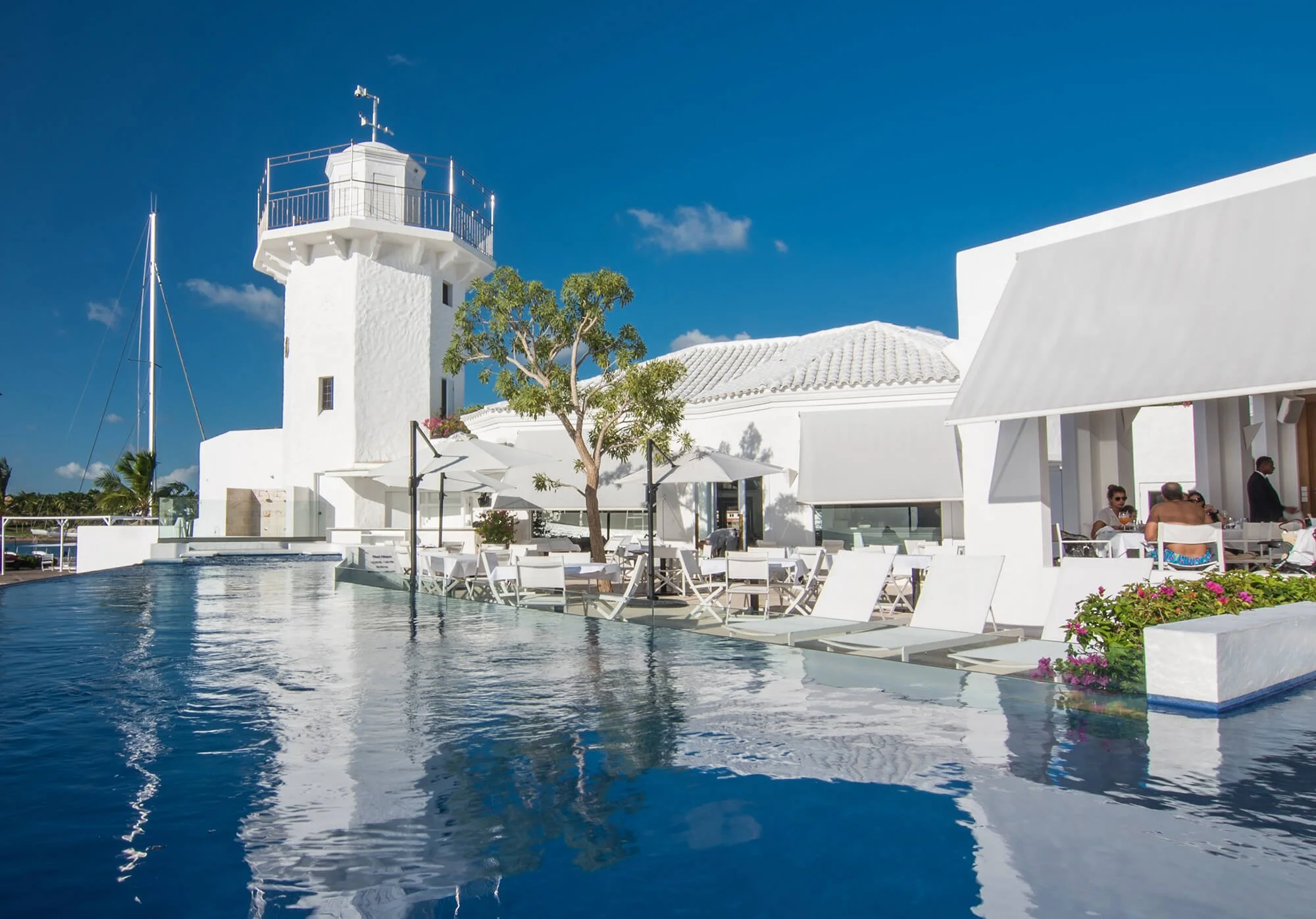 White seaside resort buildings with patio dining, a swimming pool, and a lighthouse under a clear blue sky.