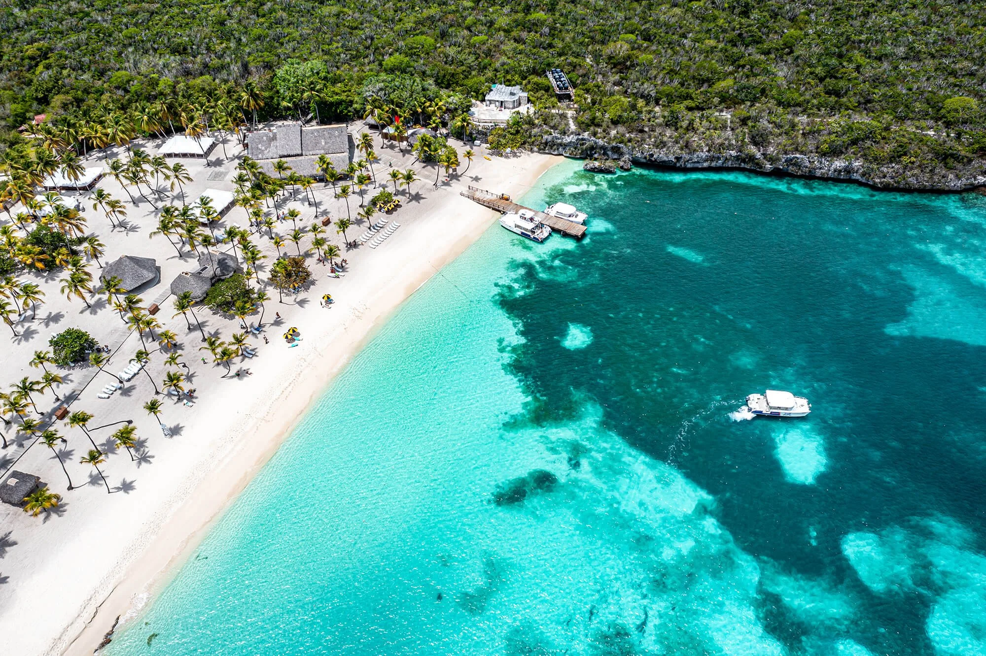 An aerial view of a tropical beach with white sand, palm trees, and clear turquoise waters. There are lounge chairs and umbrellas on the beach, along with buildings and a dock with boats anchored nearby, and dense green forest beyond the shoreline.