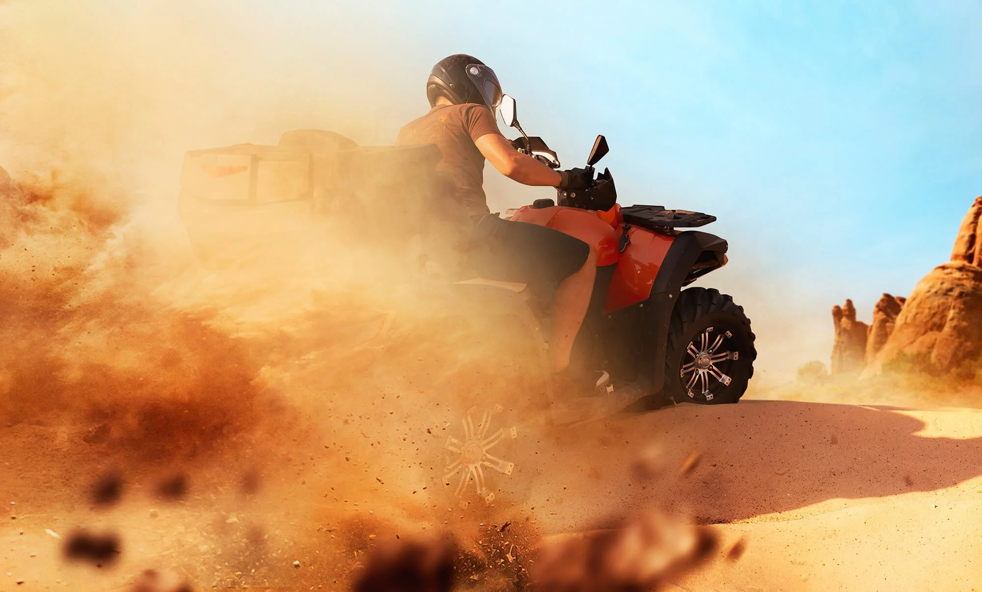 Person riding an orange all-terrain vehicle (ATV) through a desert, kicking up dirt and dust, with rocky formations in the background and clear blue sky.