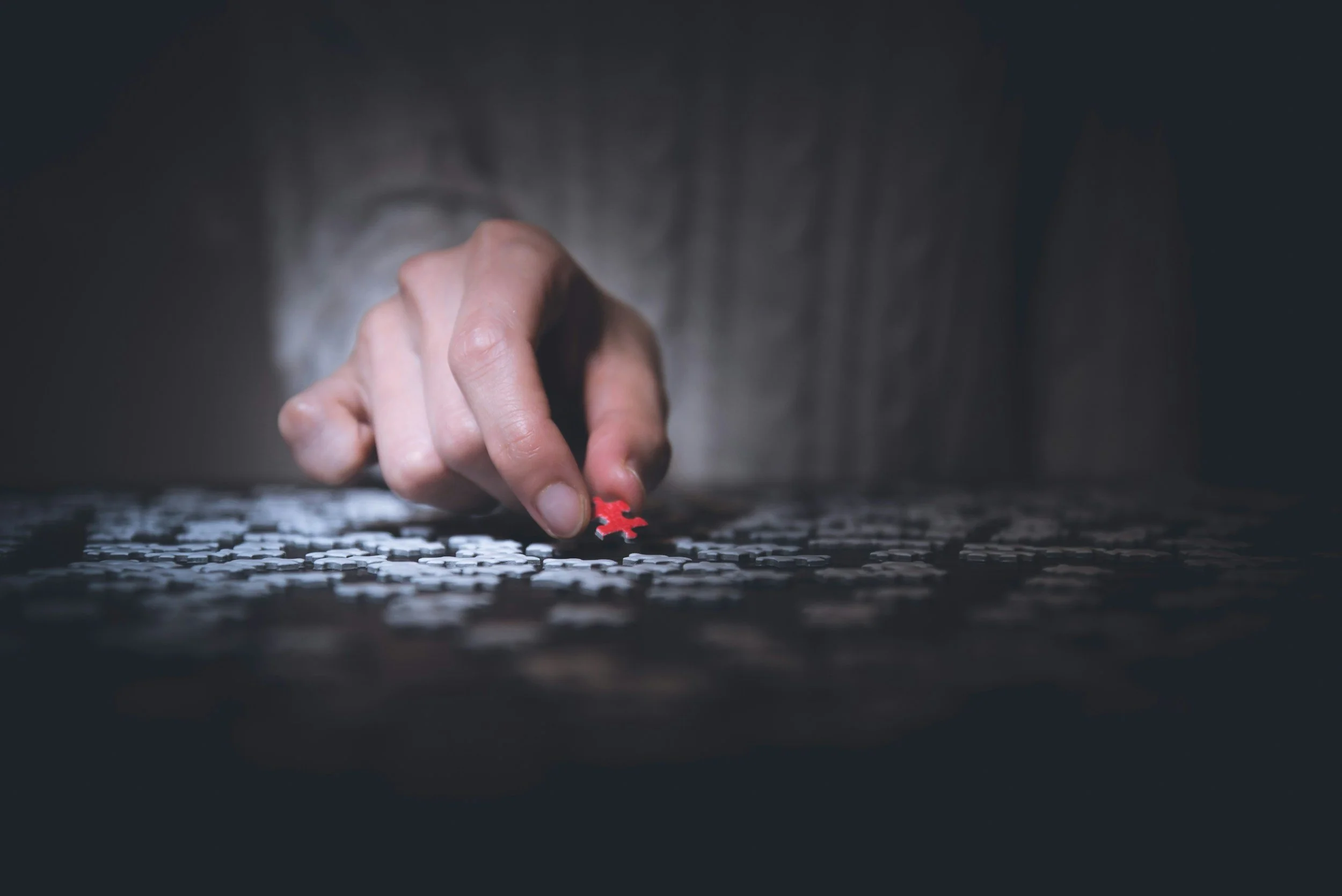 Hand holding one red puzzle piece over a table with many unconnected white puzzle pieces.