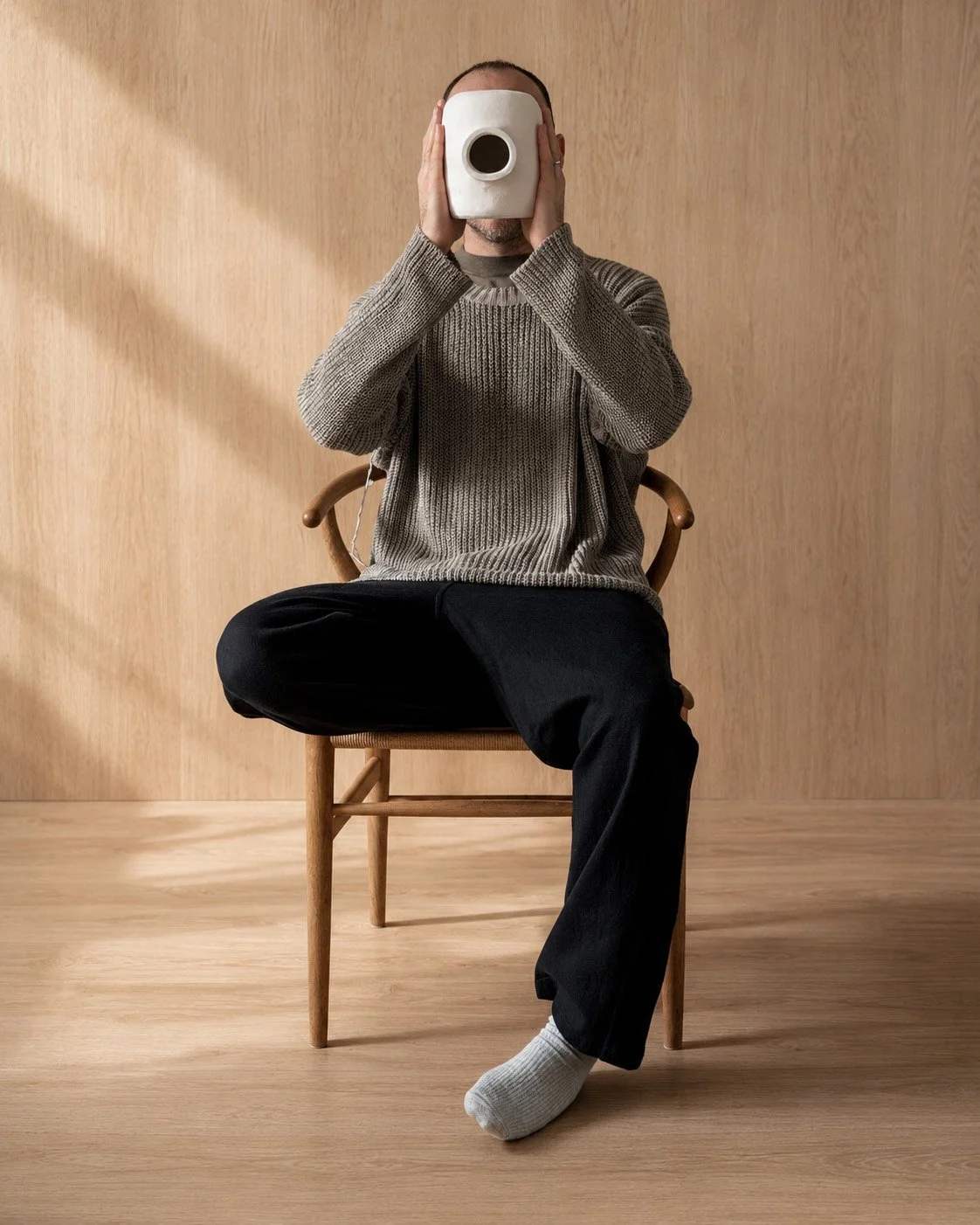 Seated Martin Lenclos holding the Jacob Vase, a ceramic object over the face in a minimal wooden room, evoking perception, identity, and design as inquiry.