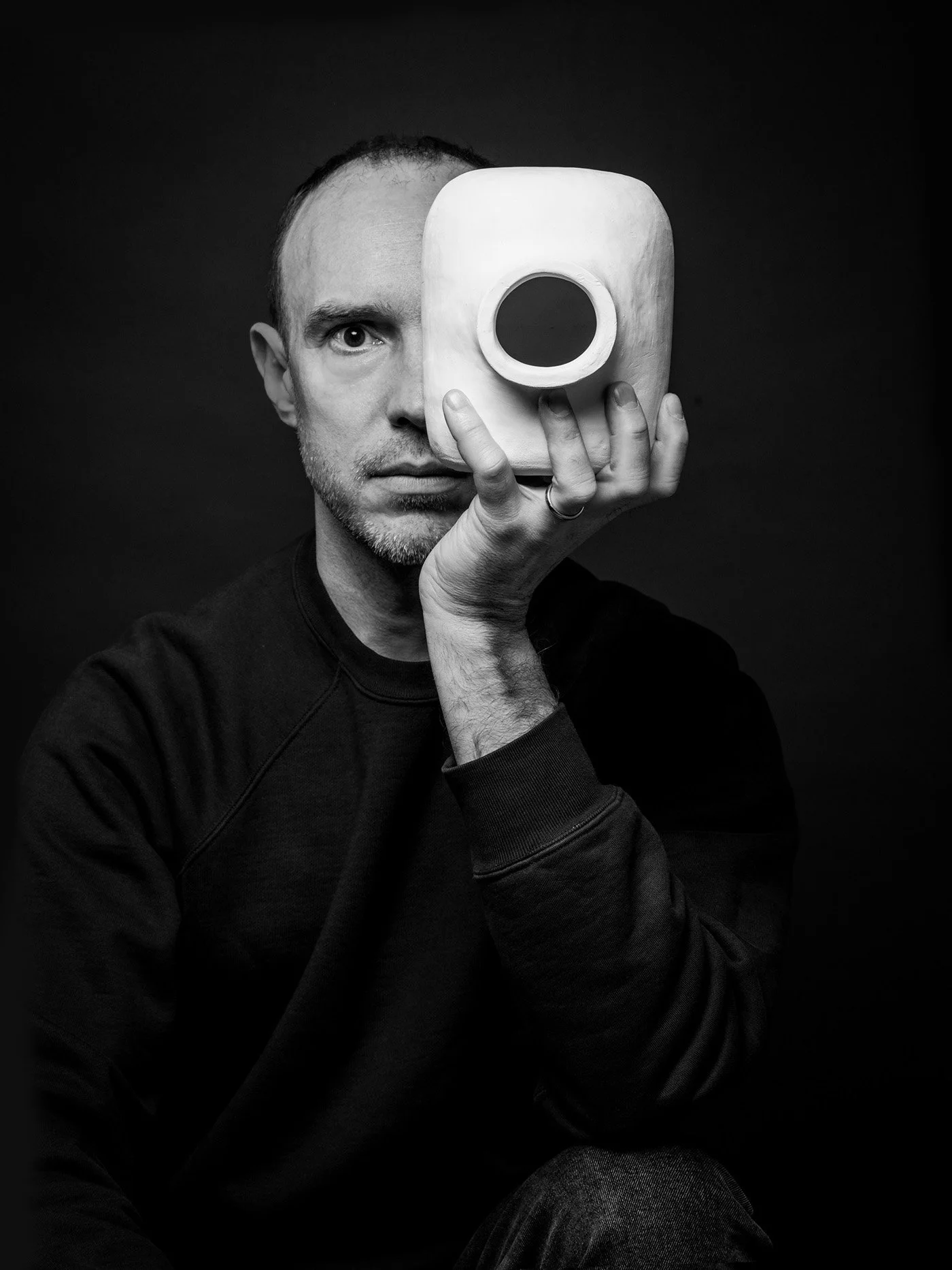 Seated Martin Lenclos holding the Jacob Vase, a ceramic object over the face in a minimal wooden room, evoking perception, identity, and design as inquiry.