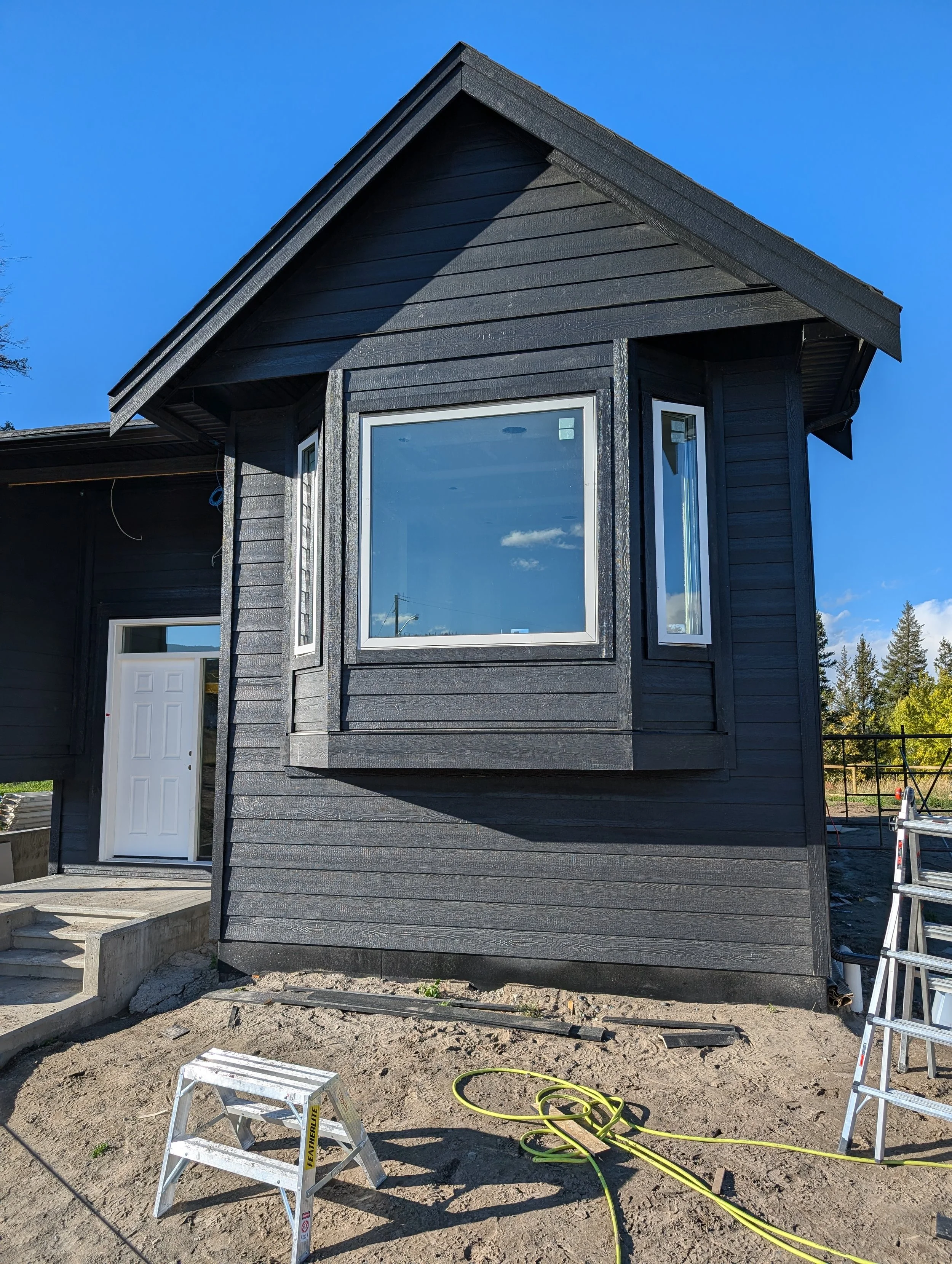 A house under construction with dark exterior siding, multiple windows, an unfinished porch area, and a saw on a stand. Professional siding installation, replacement, and exterior renovations in Williams Lake and the Cariboo Region. 