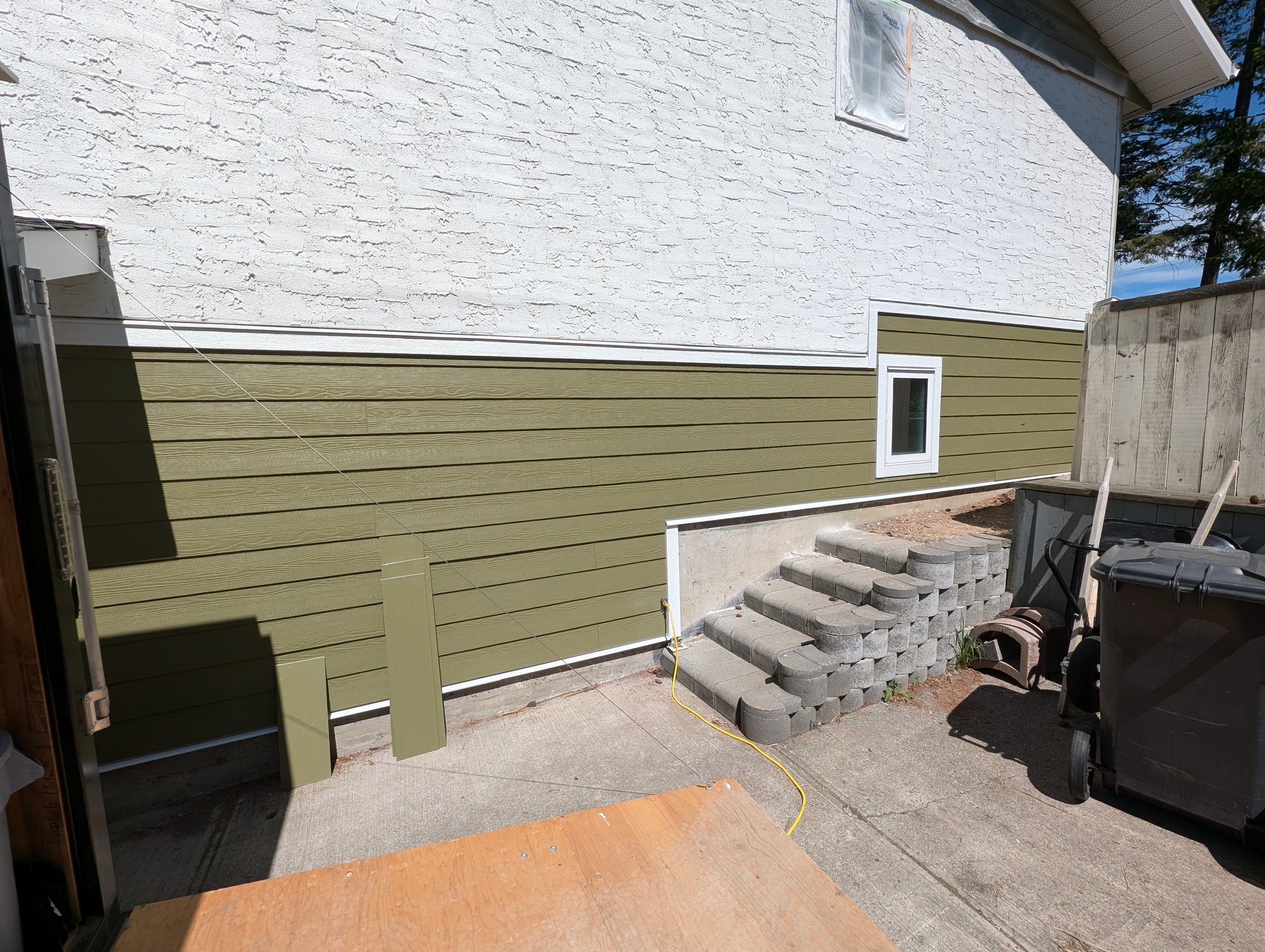 View from inside a house showing a backyard with stairs made of concrete blocks, a green house wall, and a white exterior wall of the house with small rectangular window.