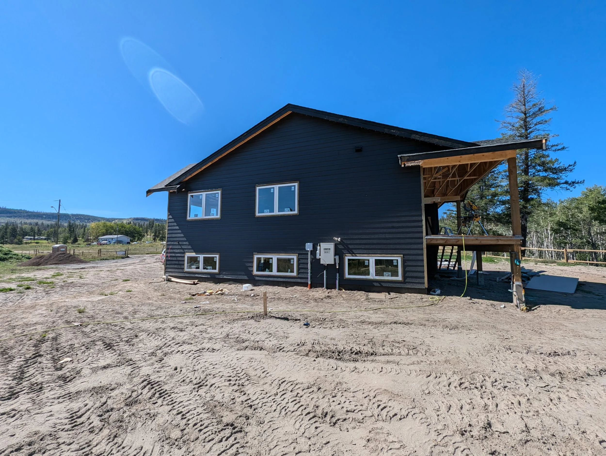 A house under construction with dark exterior siding, multiple windows, an unfinished porch area, and a saw on a stand. Professional siding installation, replacement, and exterior renovations in Williams Lake and the Cariboo Region. 