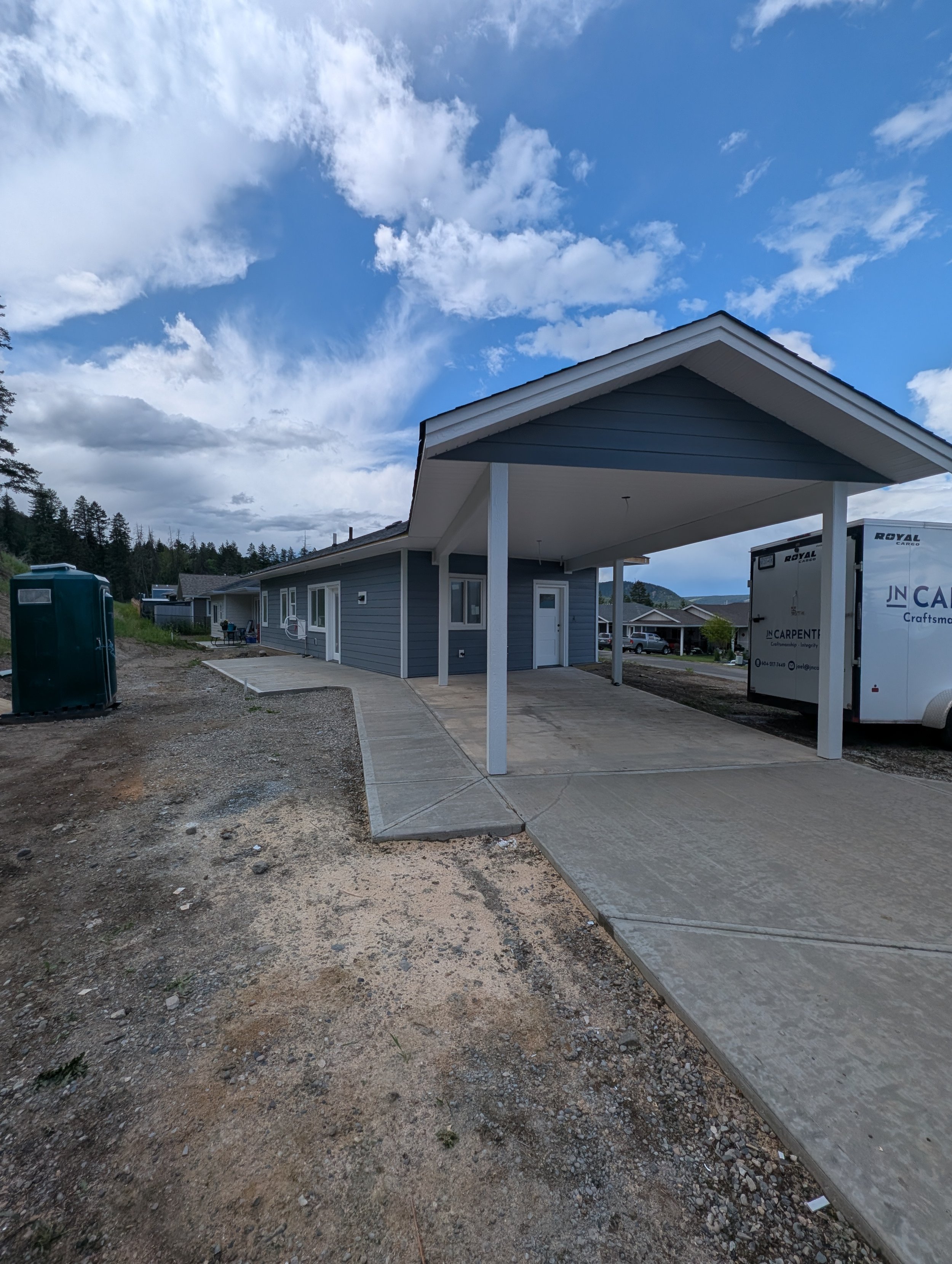A house under construction with blue siding and a dark gray roof, in front of a wooded area with tall trees and a cloudy sky. Professional siding installation, replacement, and exterior renovations in Williams Lake and the Cariboo Region. 