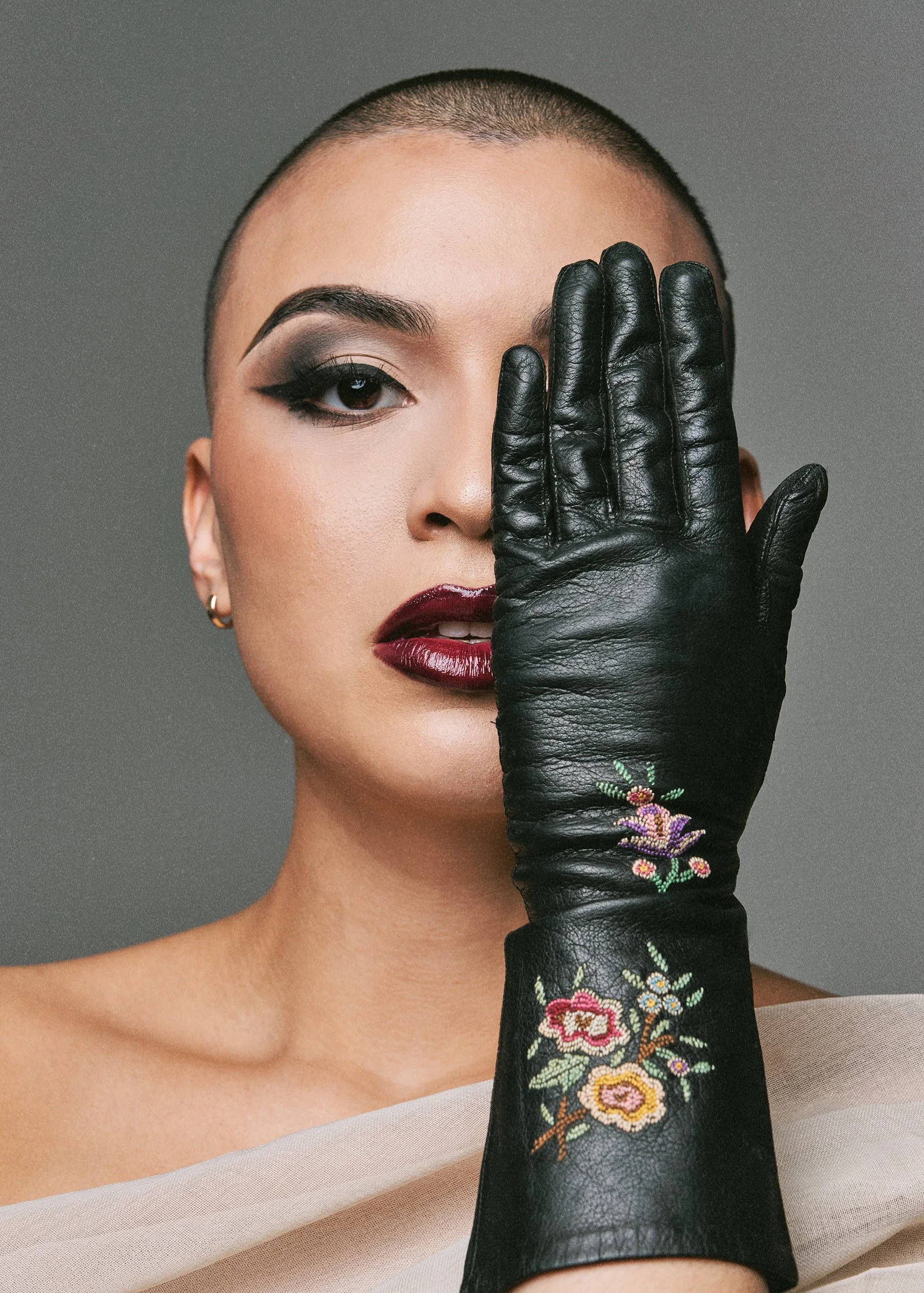 A woman with short hair, bold makeup, and earrings covering one eye with a black leather glove embroidered with colorful flowers.
