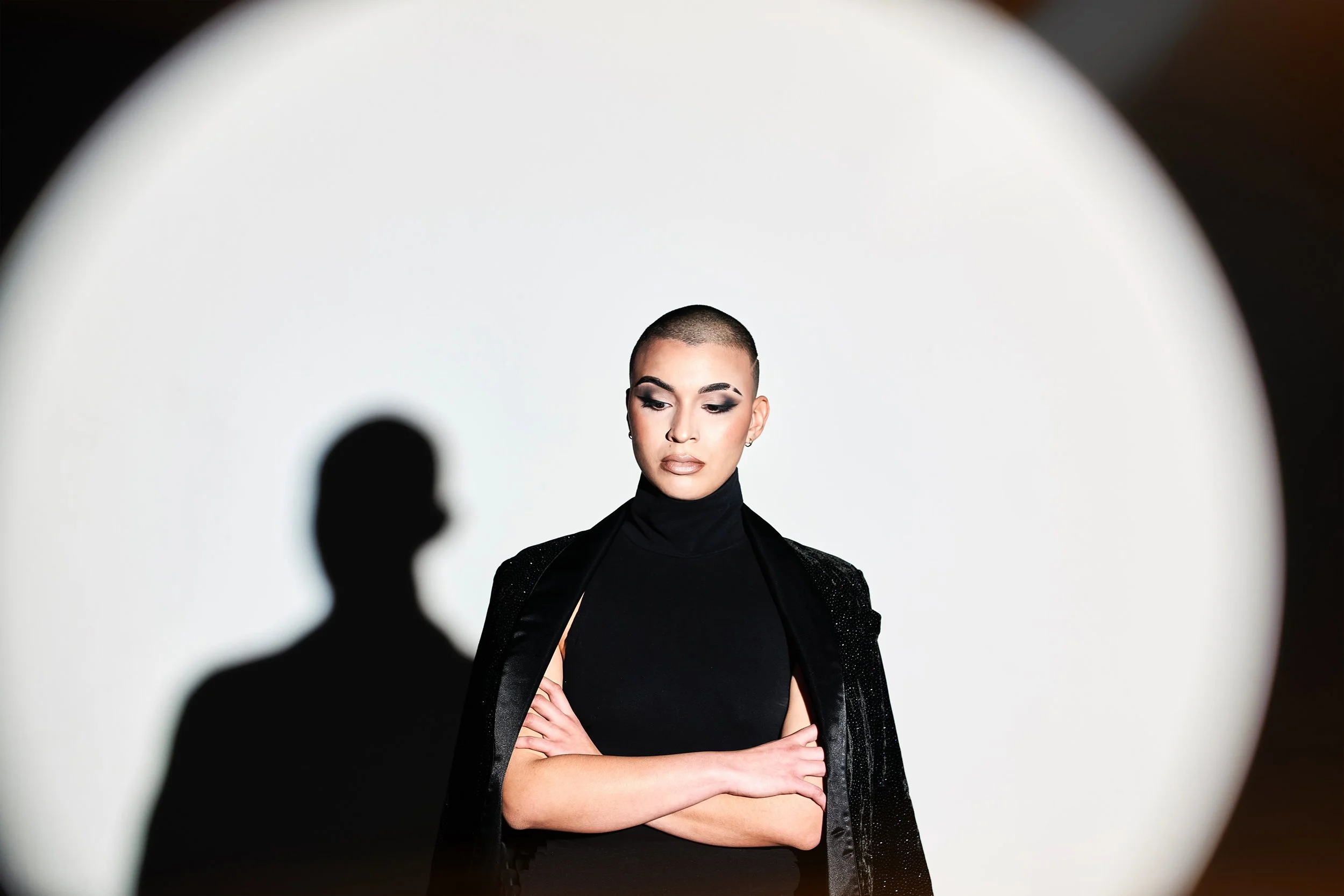A woman with a shaved head, wearing a black turtleneck and a black jacket, poses with her arms crossed against a plain background.