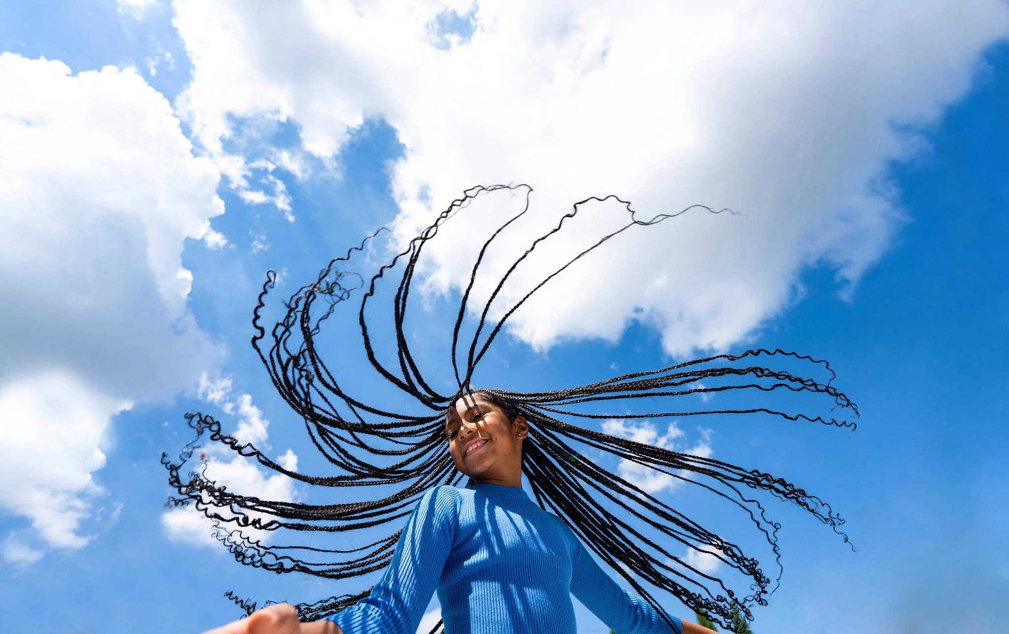 Woman with long braided hair wearing a blue sweater standing against a blue sky with clouds.