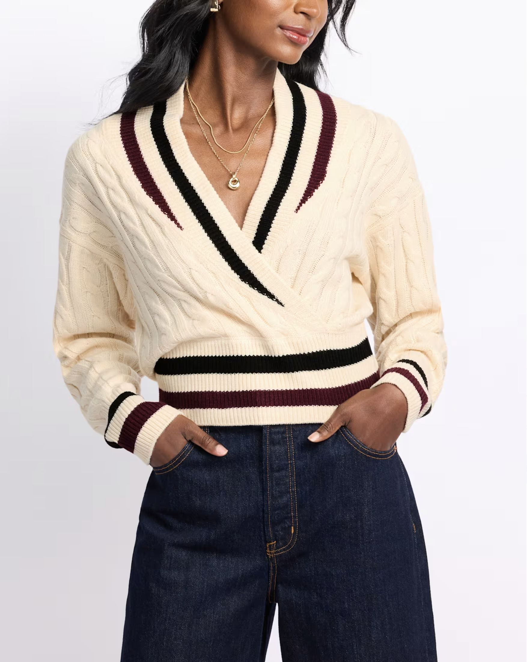 A woman wearing a cream-colored V-neck sweater with black, burgundy, and cream stripes, paired with dark blue jeans and layered gold necklaces.
