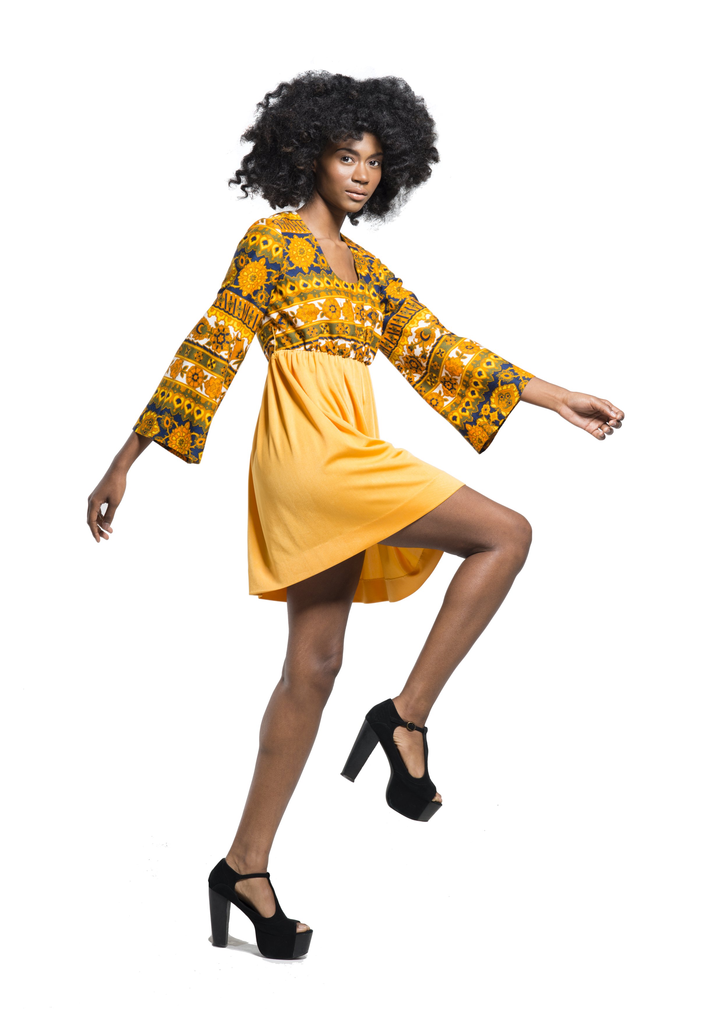 A woman with curly black hair, wearing a colorful ethnic patterned top with yellow, orange, and blue, and a yellow skirt, standing with one leg raised and arms extended, against a white background.