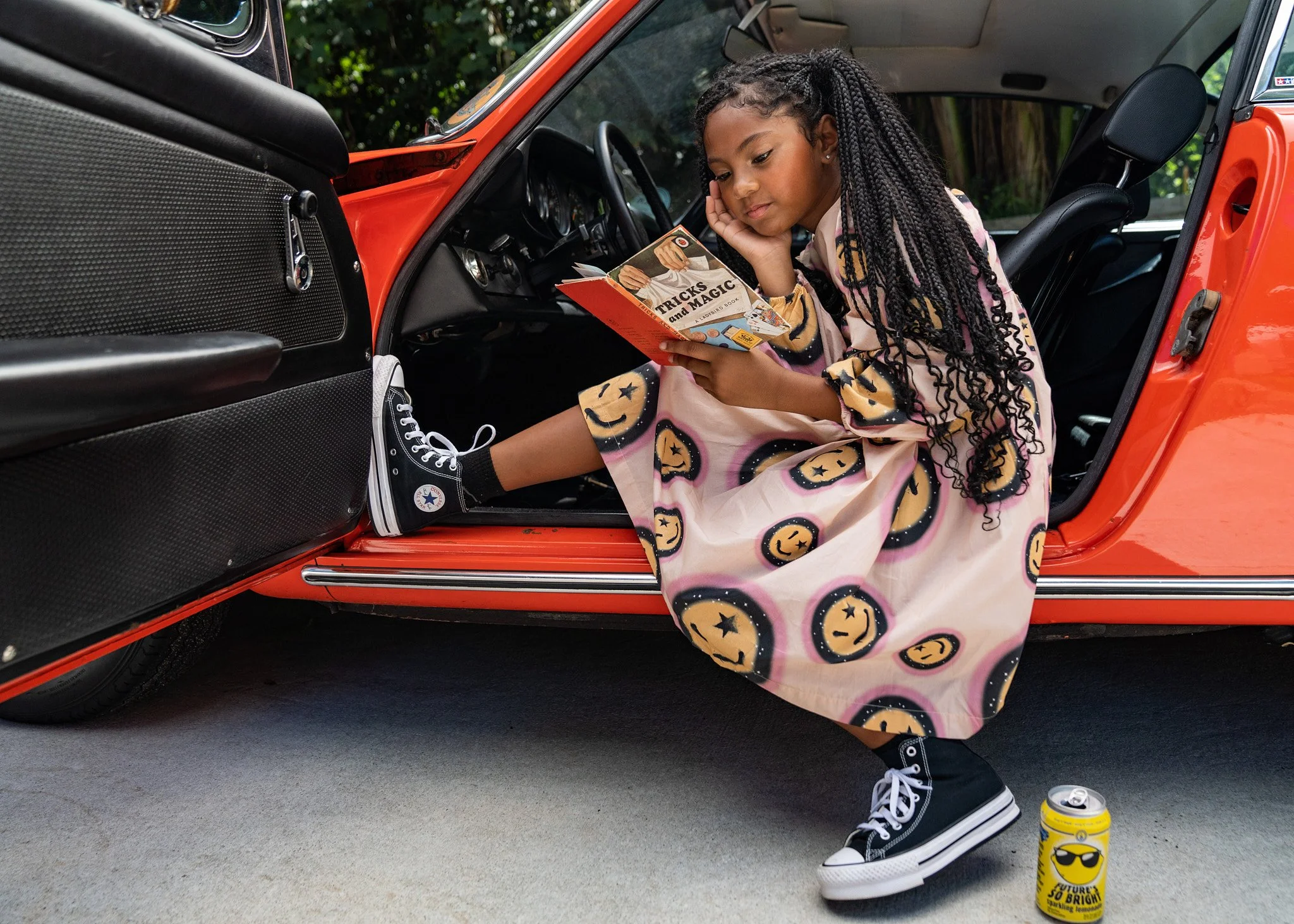 A young girl with braided hair, wearing a pink dress with orange and black smiley face patterns and black high-top Converse sneakers, sitting in the driver's seat of an orange vintage car. She is reading a book titled "Tricks and Magic," with her lef