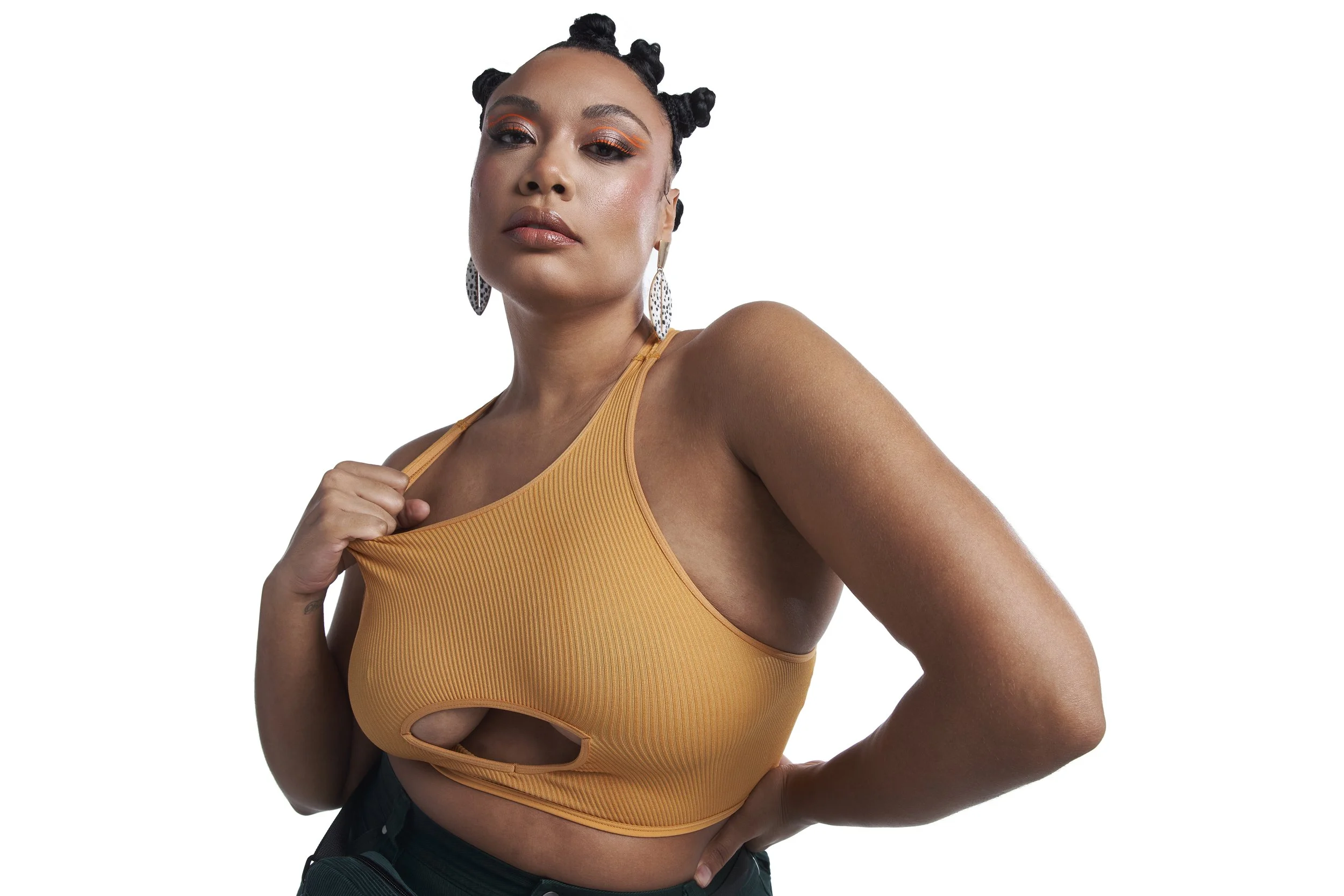 A woman with dark hair styled in bantu knots, wearing orange eyeshadow and earrings, a yellow sleeveless top with a cutout, and dark pants, posing with her hand on her hip against a white background.