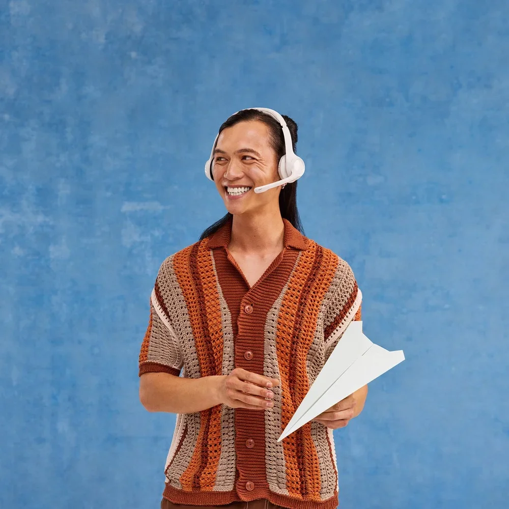 A person wearing a colorful striped crochet cardigan holding papers and smiling, wearing a white headset with a microphone, against a blue background.