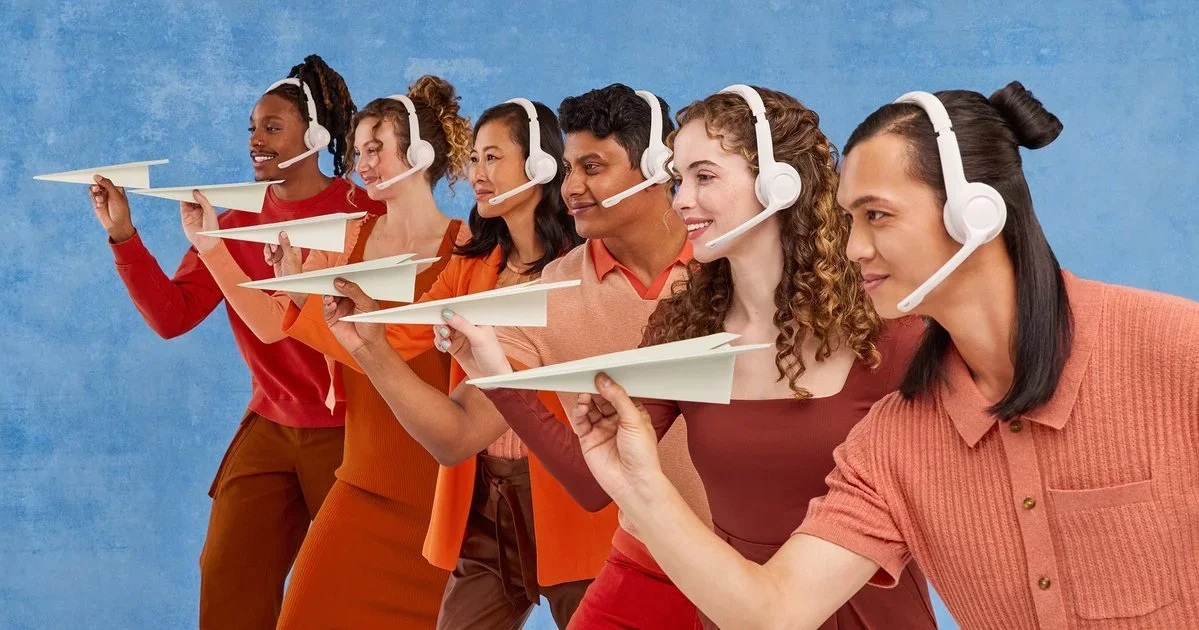 Six diverse people wearing headsets with microphones, holding paper airplane models, and smiling against a blue background.