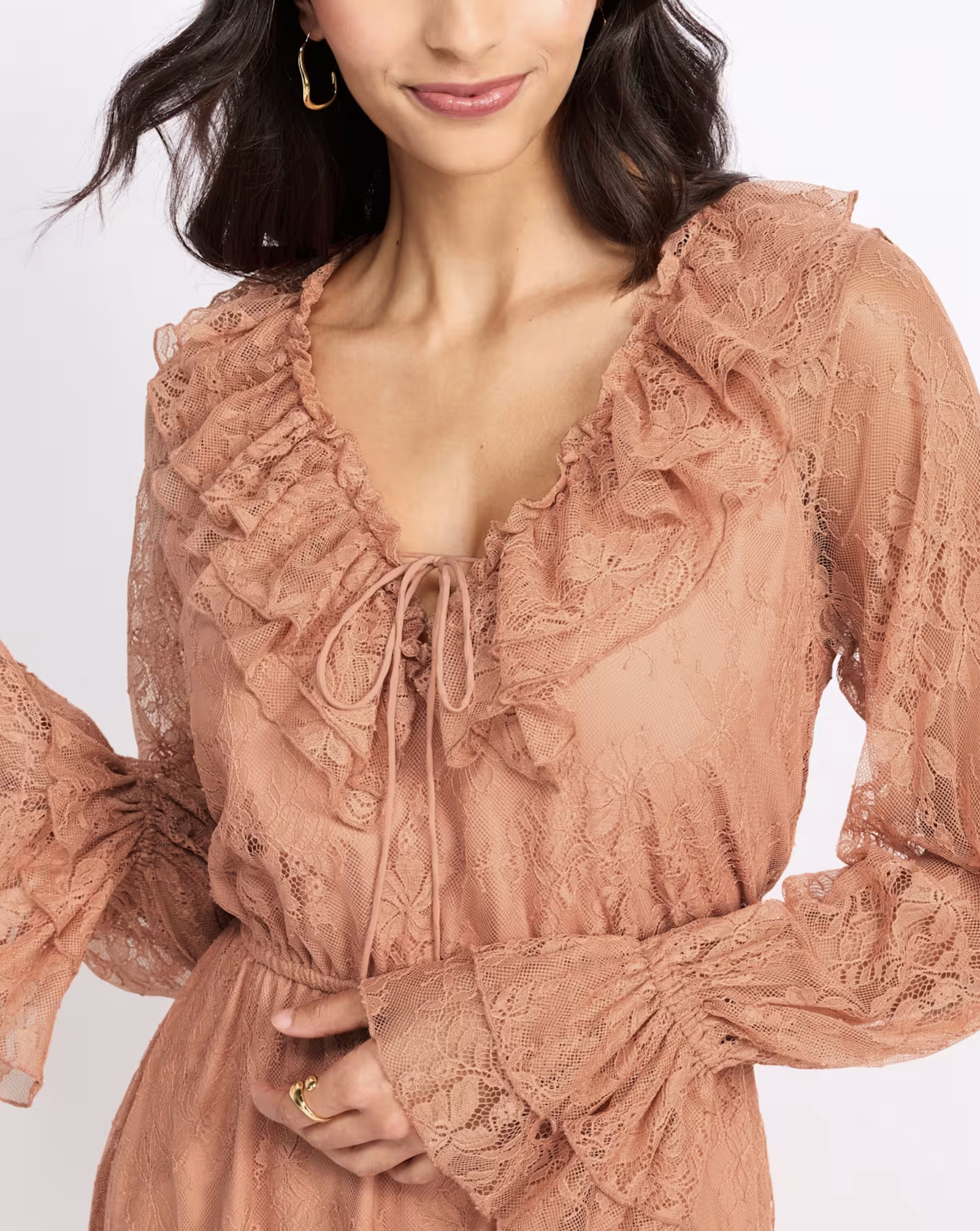 A woman with dark hair and gold hoop earrings, smiling slightly, wearing a peach-colored lace dress with ruffles and a tie at the neckline.