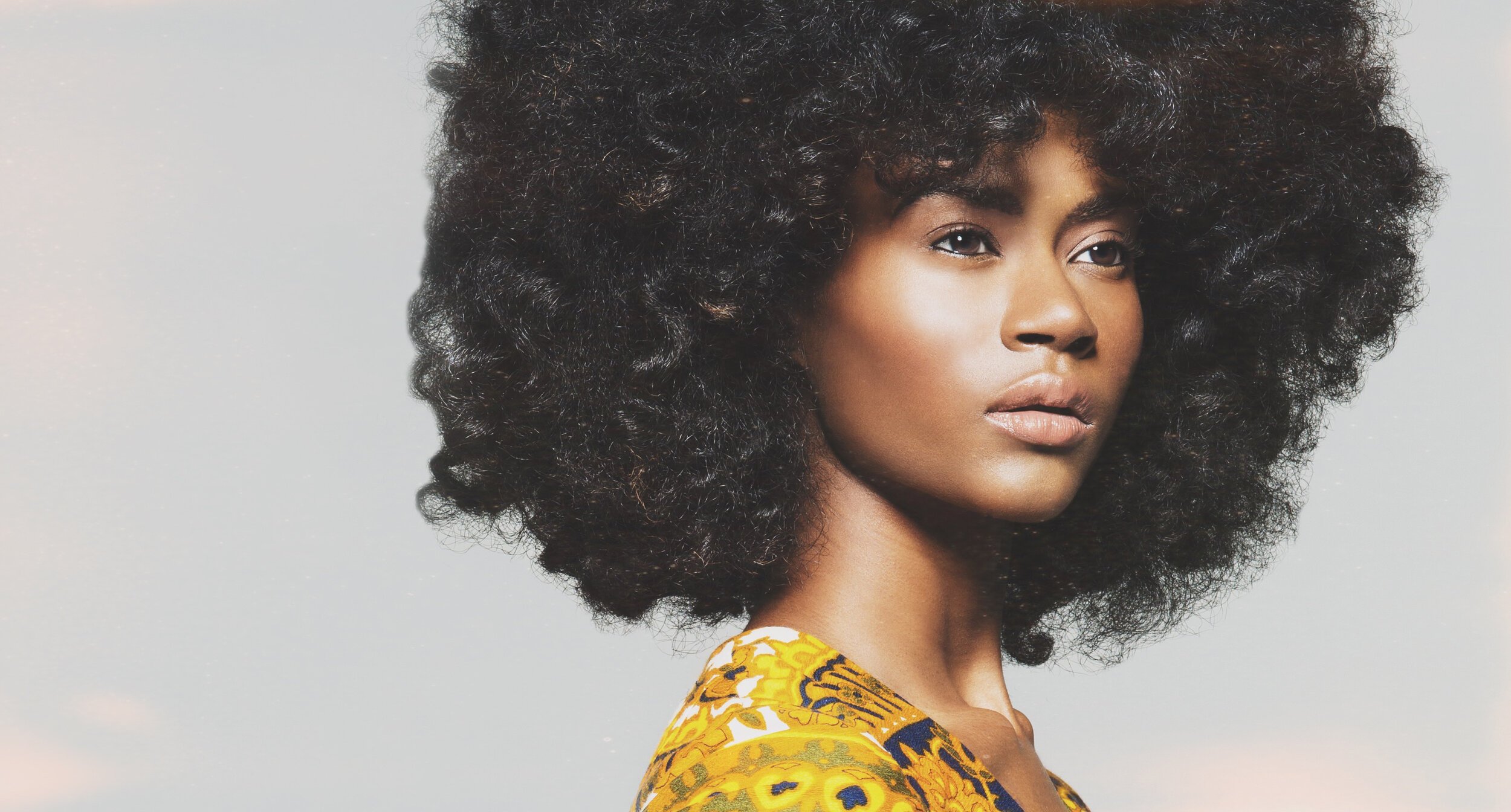 A woman with voluminous curly black hair and smooth skin, wearing a yellow patterned top, against a neutral background.