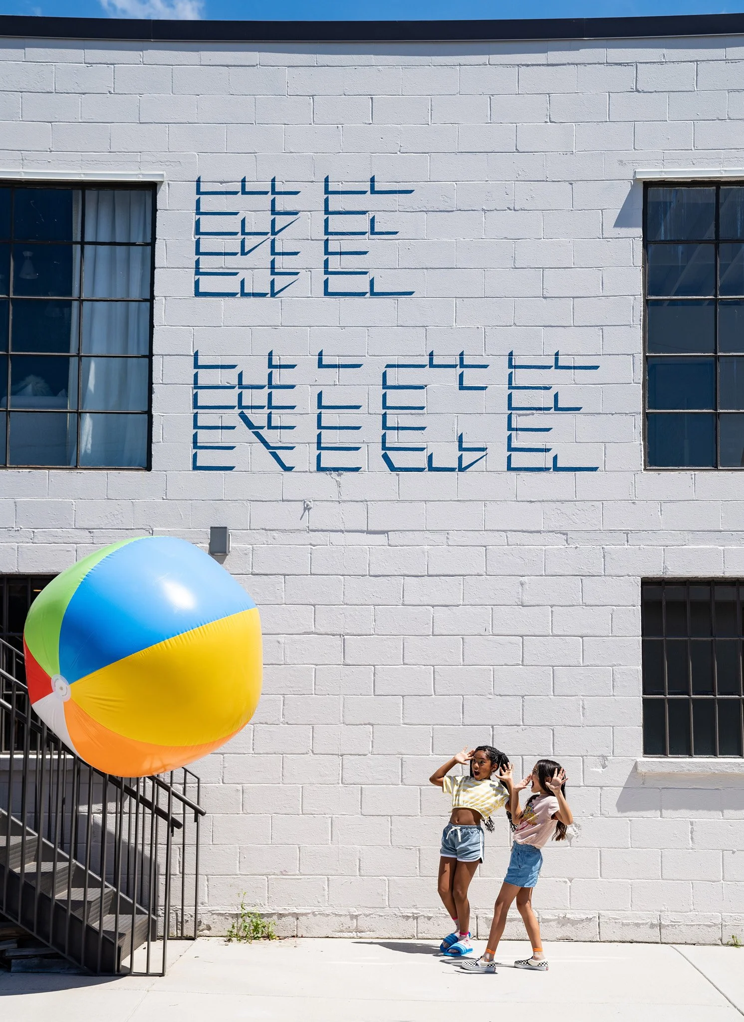 Two girls standing in front of a white brick wall with a colorful beach ball nearby. The wall has a sign that says 'Cool Teen' in blue letters. The girls are wearing casual summer clothes, and the scene appears to be outdoors on a sunny day.