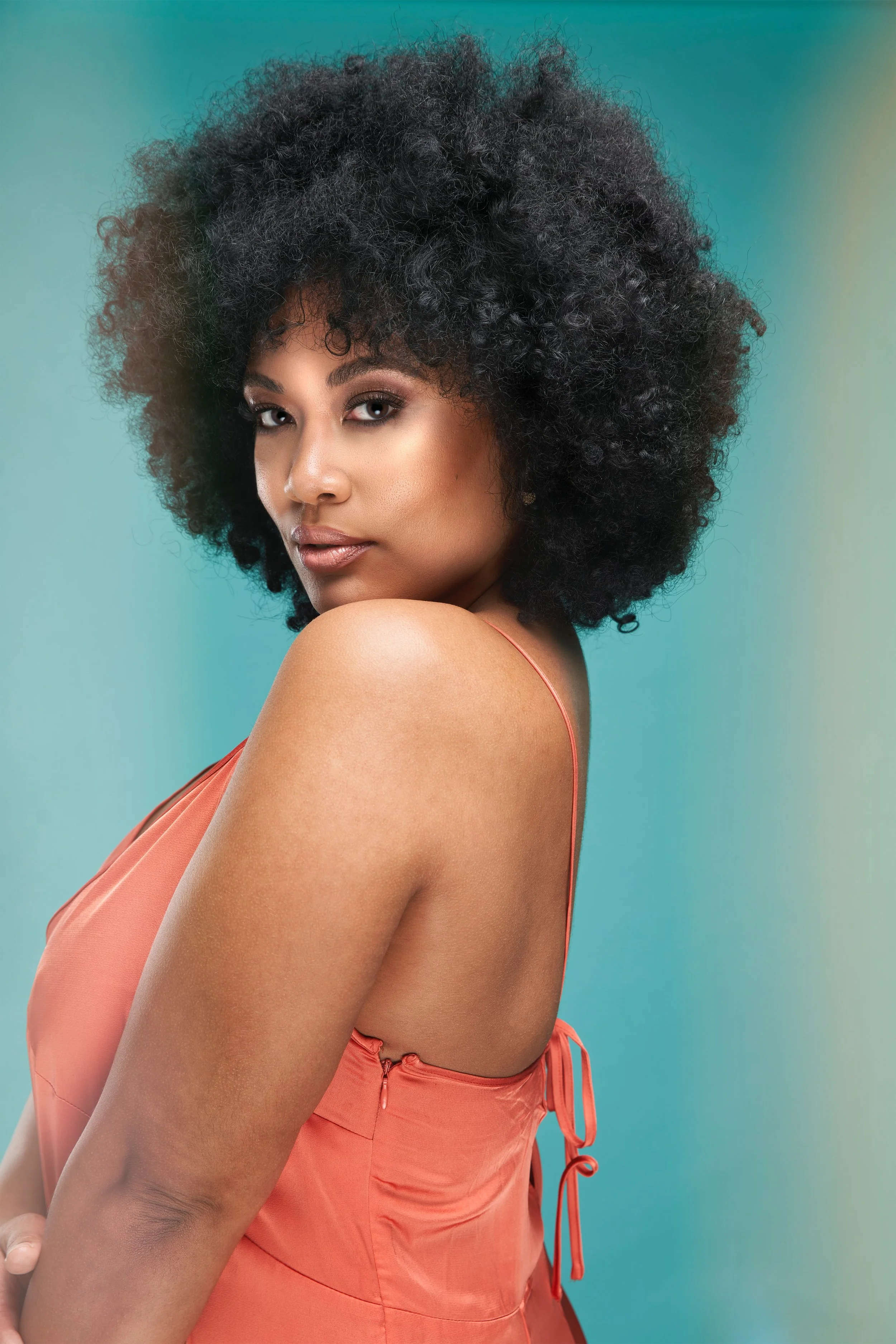 A woman with dark curly hair, wearing a sleeveless peach-colored dress, posing against a gradient background from teal to yellow.