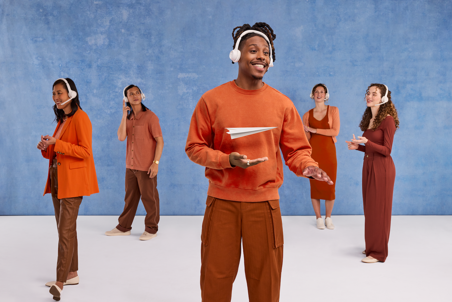 Group of five diverse young adults wearing headphones, standing against a blue textured background, with one person in the foreground holding a paper airplane.