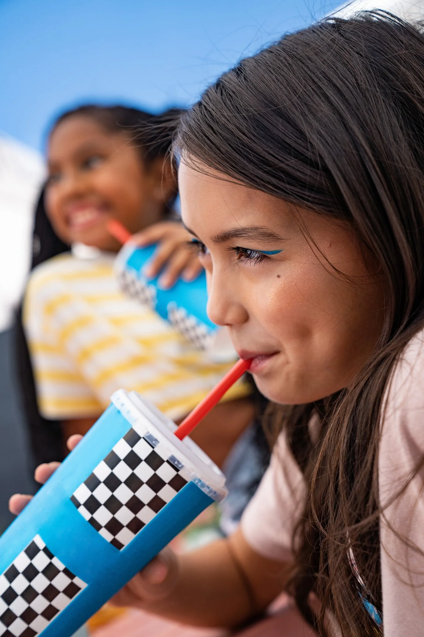 Two girls sitting in a car, drinking from large cups with checkered patterns, enjoying their drinks.