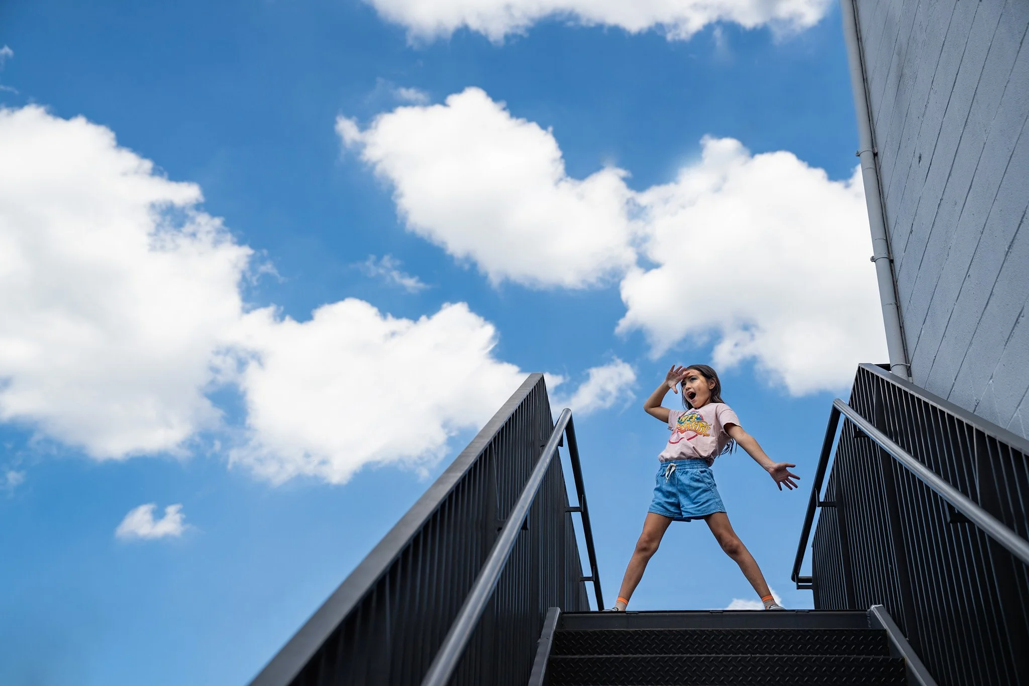 Young girl on outdoor staircase, standing with legs apart, hand on forehead as if looking into the distance, sky with scattered clouds in background.