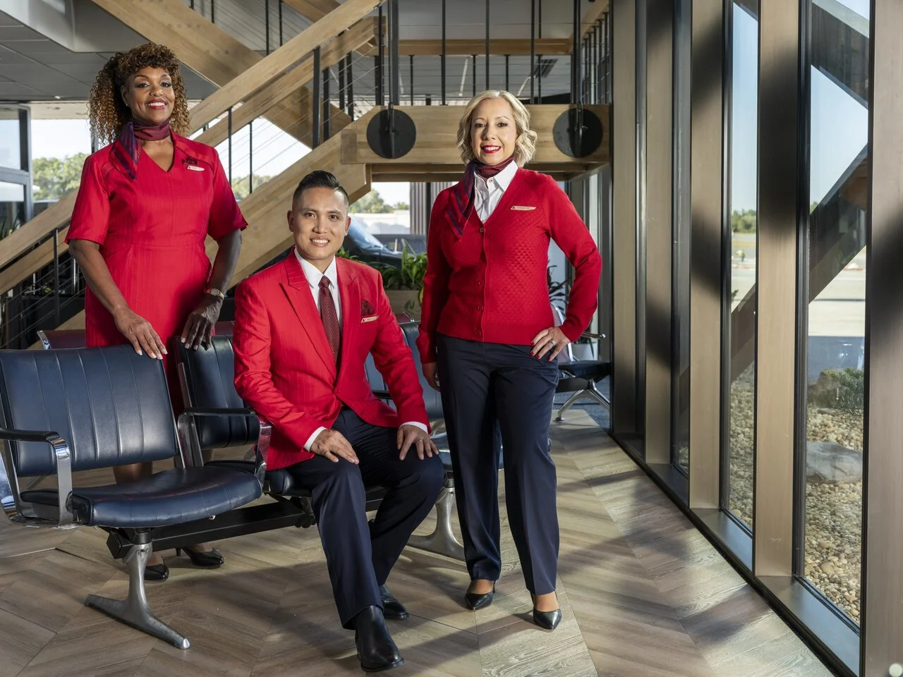 Three employees in red uniforms inside an airport or hotel lobby with large windows and stairs behind them.