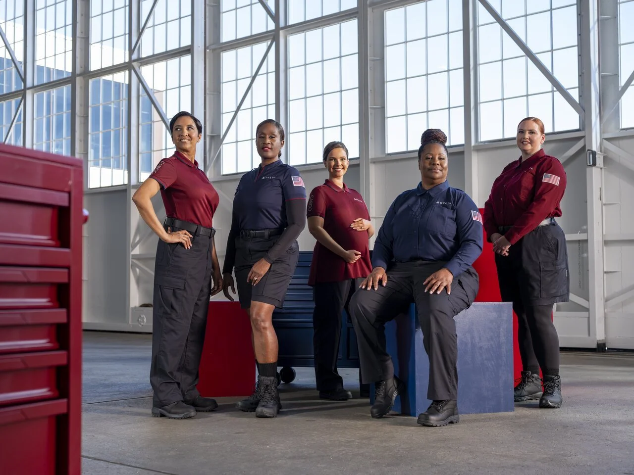 Group of five women working at a space agency or NASA, posing inside a high-ceiling hangar or workshop, dressed in NASA or space agency uniforms, with equipment and large windows in the background.