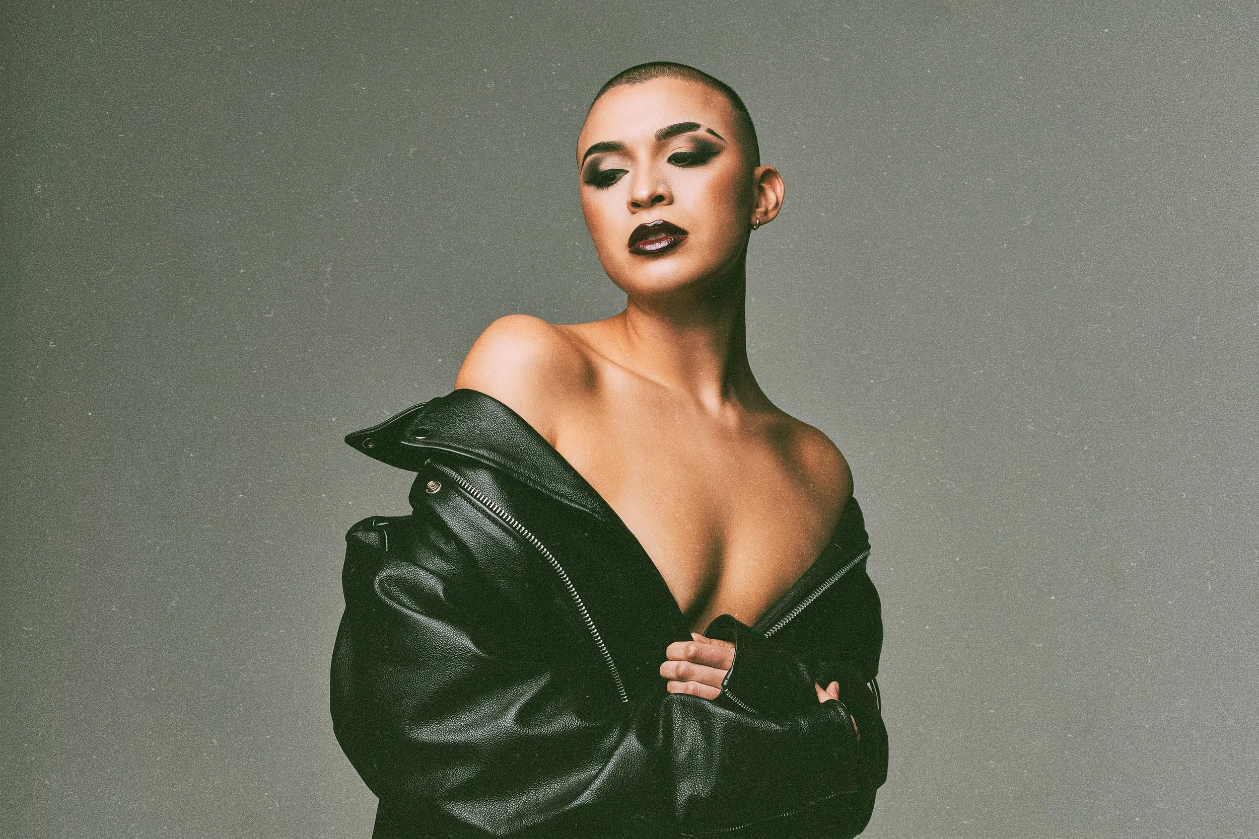 A woman with shaved head and makeup, wearing a black leather jacket off the shoulders, with a gray background.