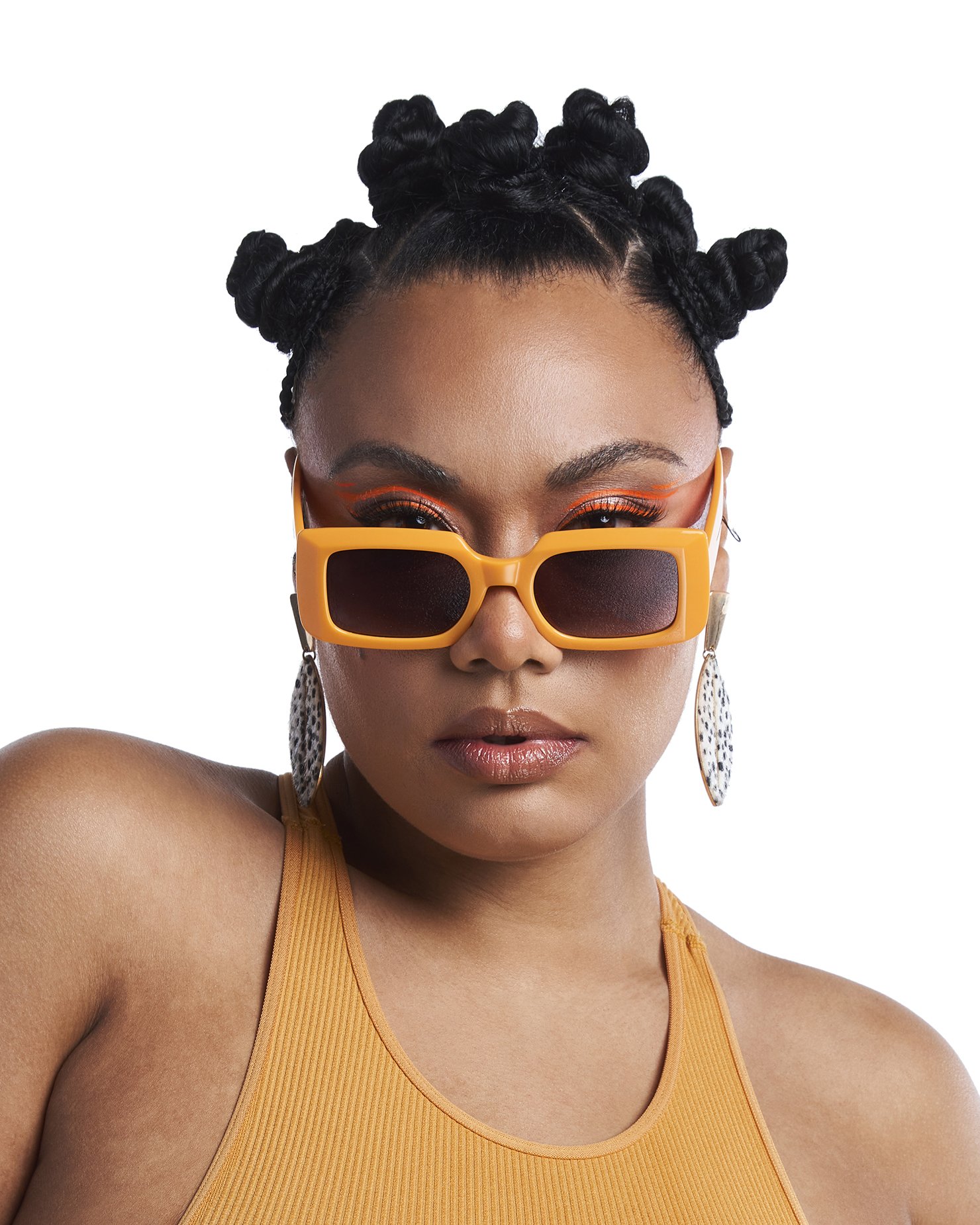 A woman with styled black hair in bantu knots, wearing rectangular orange sunglasses, large silver earrings, orange eye makeup, and a mustard tank top.