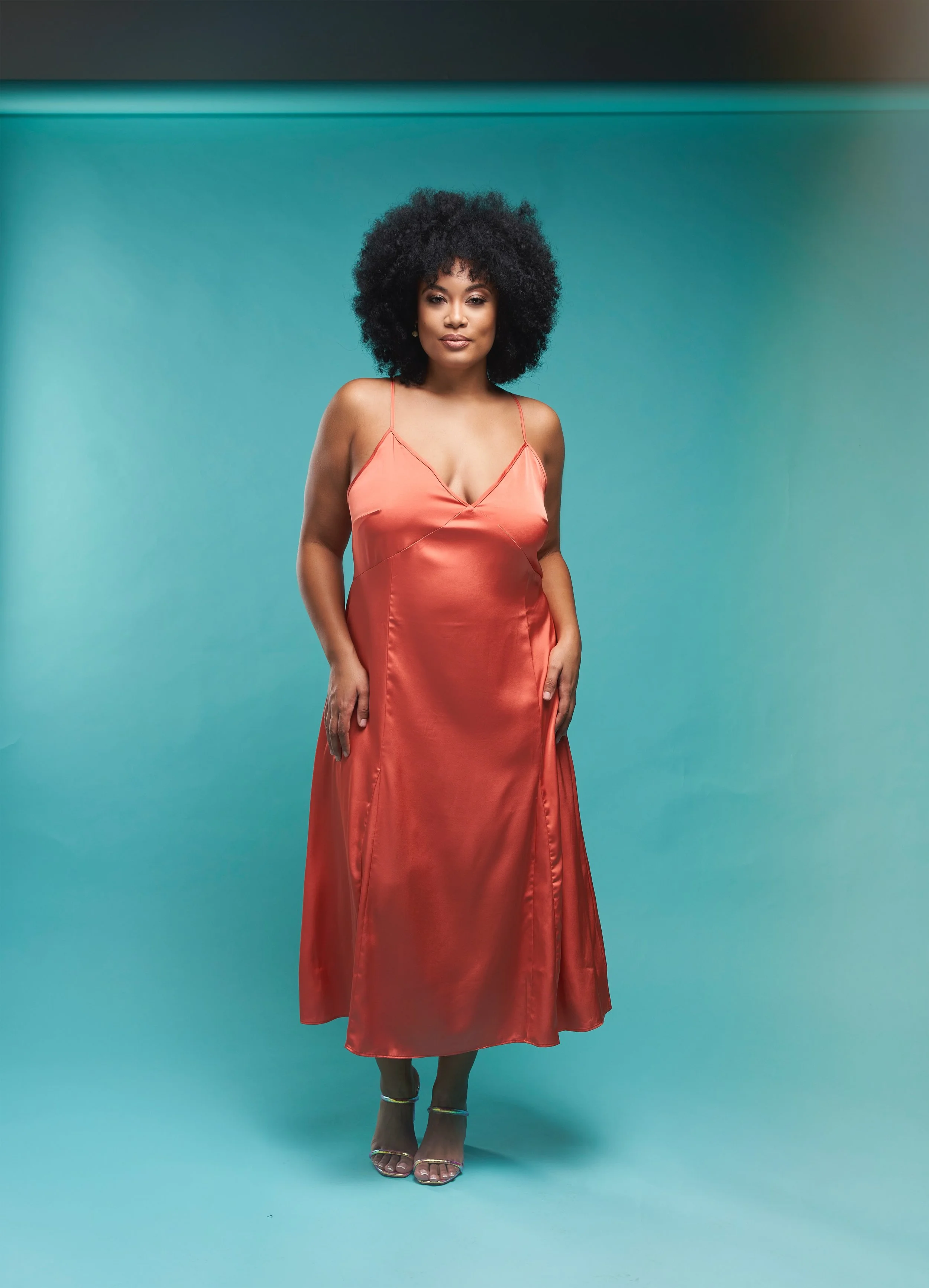 A woman with natural curly hair wearing a coral satin slip dress and silver high heels, standing against a turquoise background.