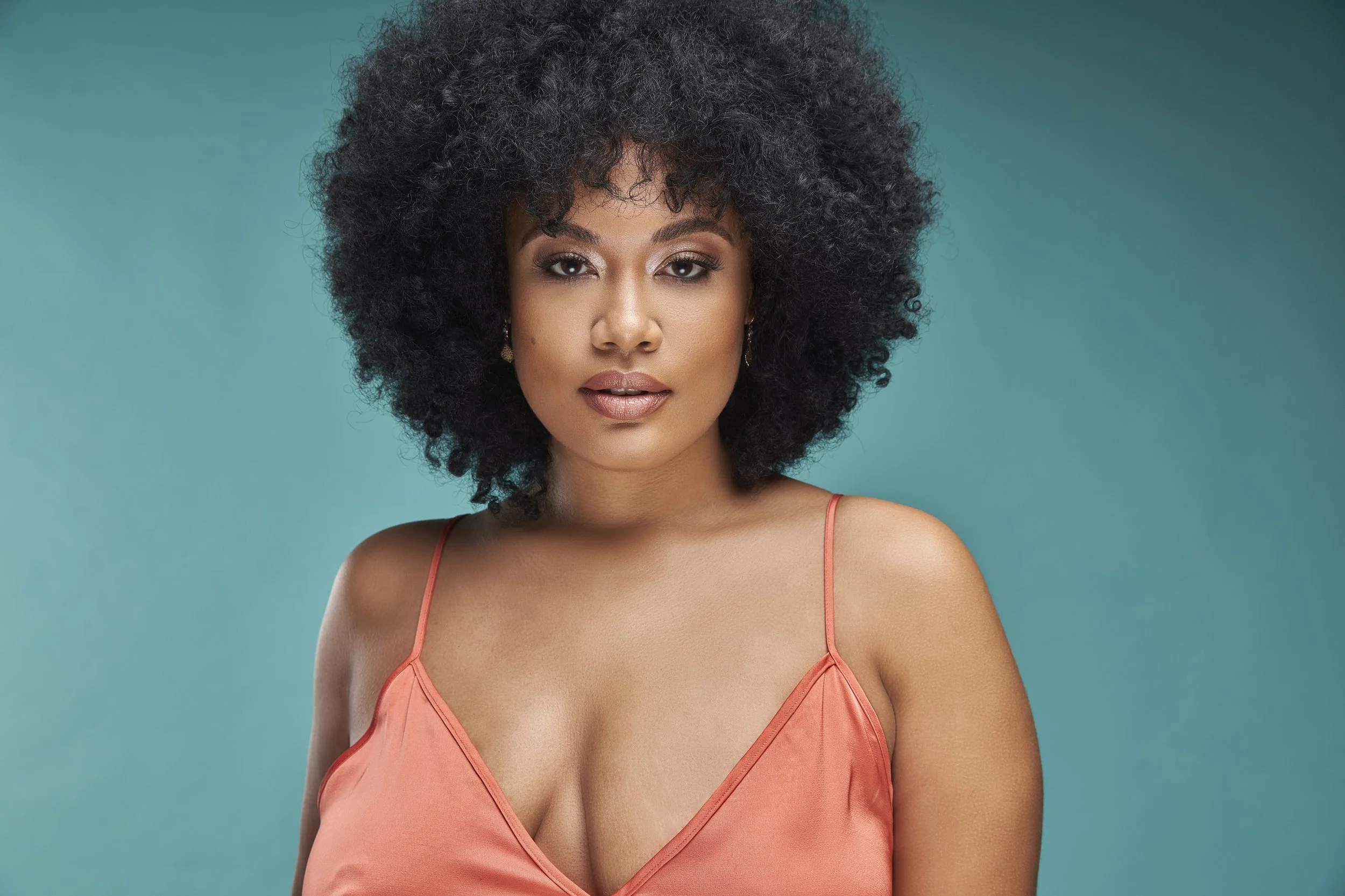 A woman with dark, curly hair wearing a coral spaghetti strap dress against a teal background.