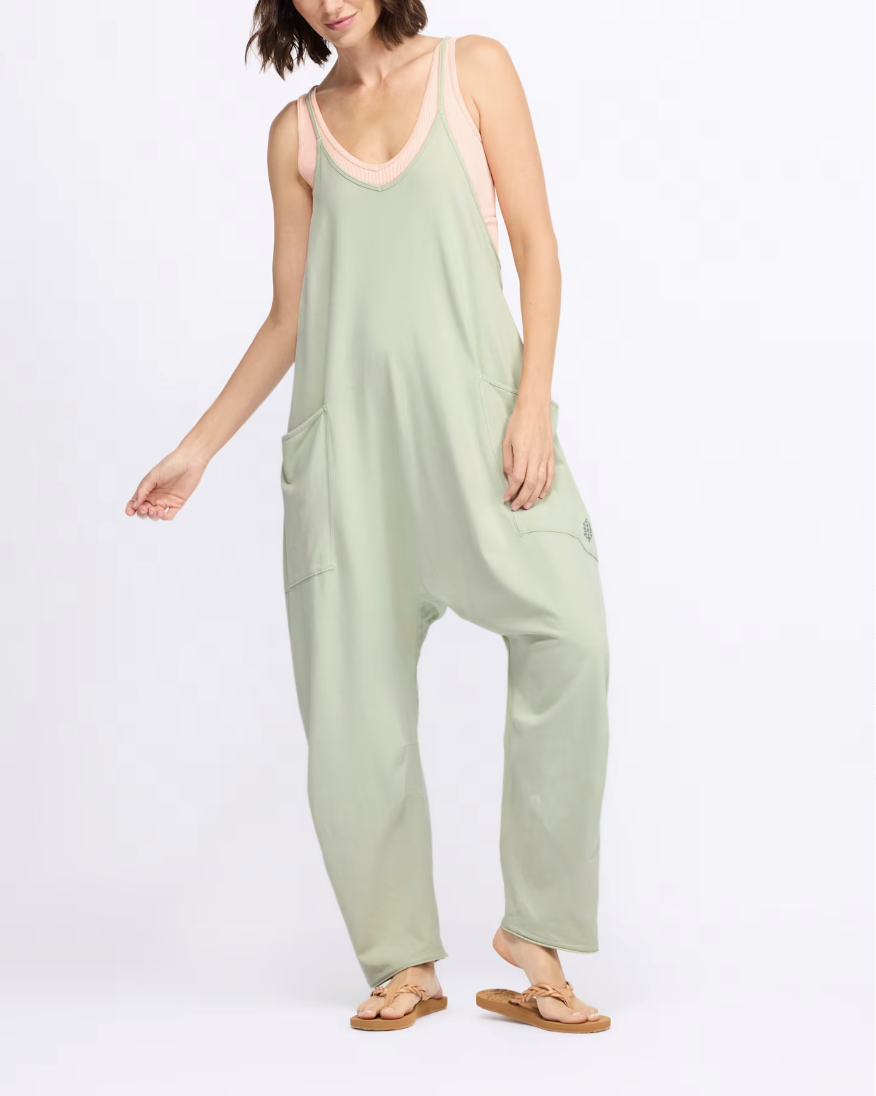 A woman wearing a pastel green jumpsuit with pockets, a pink tank top, and tan sandals, standing against a plain white background.