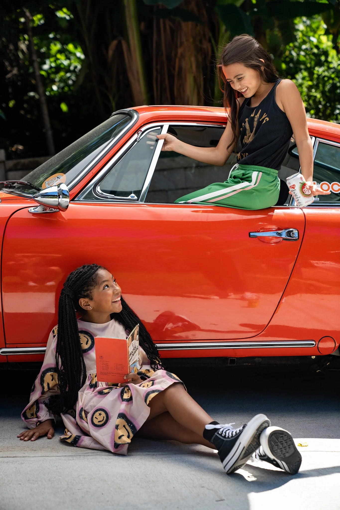 A young woman sitting in a red vintage car talking to a girl sitting on the ground outside the car, holding a snack and smiling.