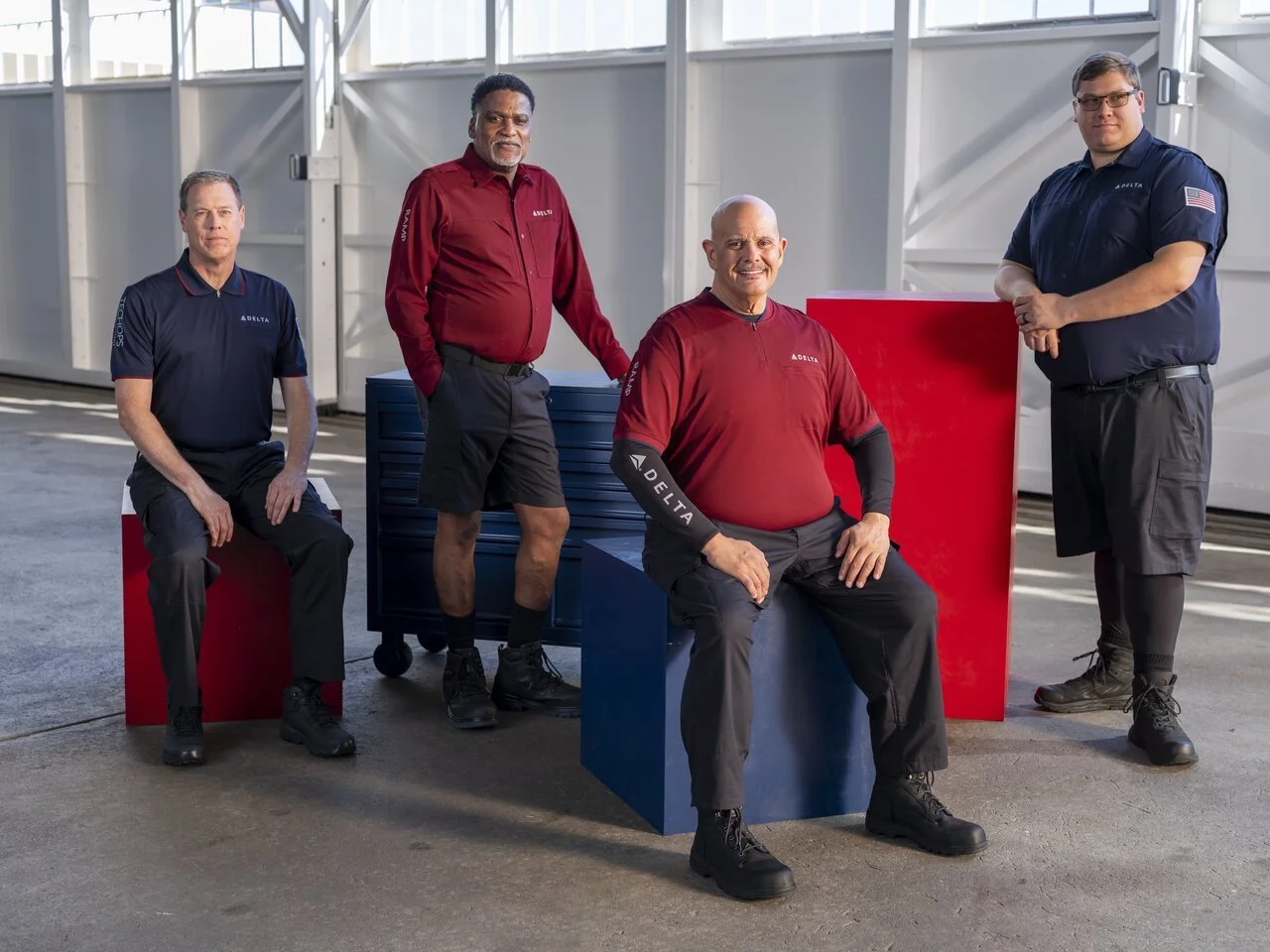 Five men in aviation uniforms posing in a hangar with storage boxes and geometric structures in the background.