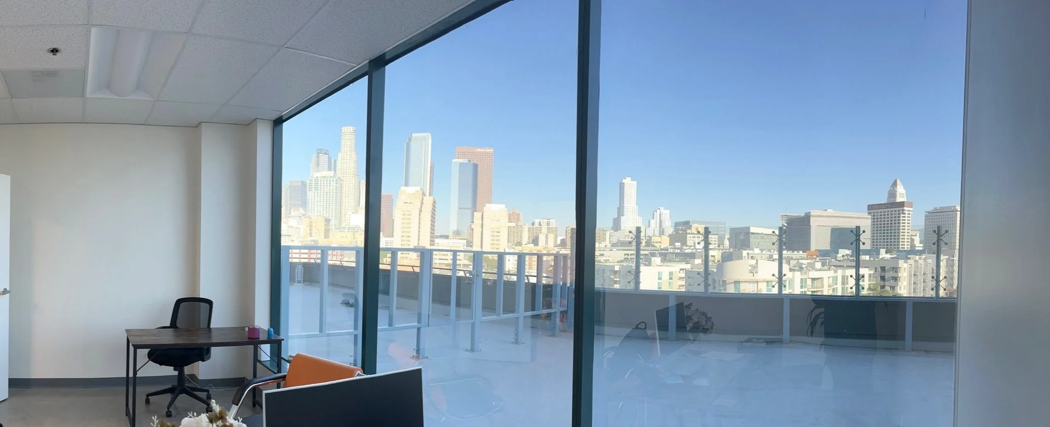 Office interior with large windows overlooking a cityscape of tall buildings under a clear blue sky.