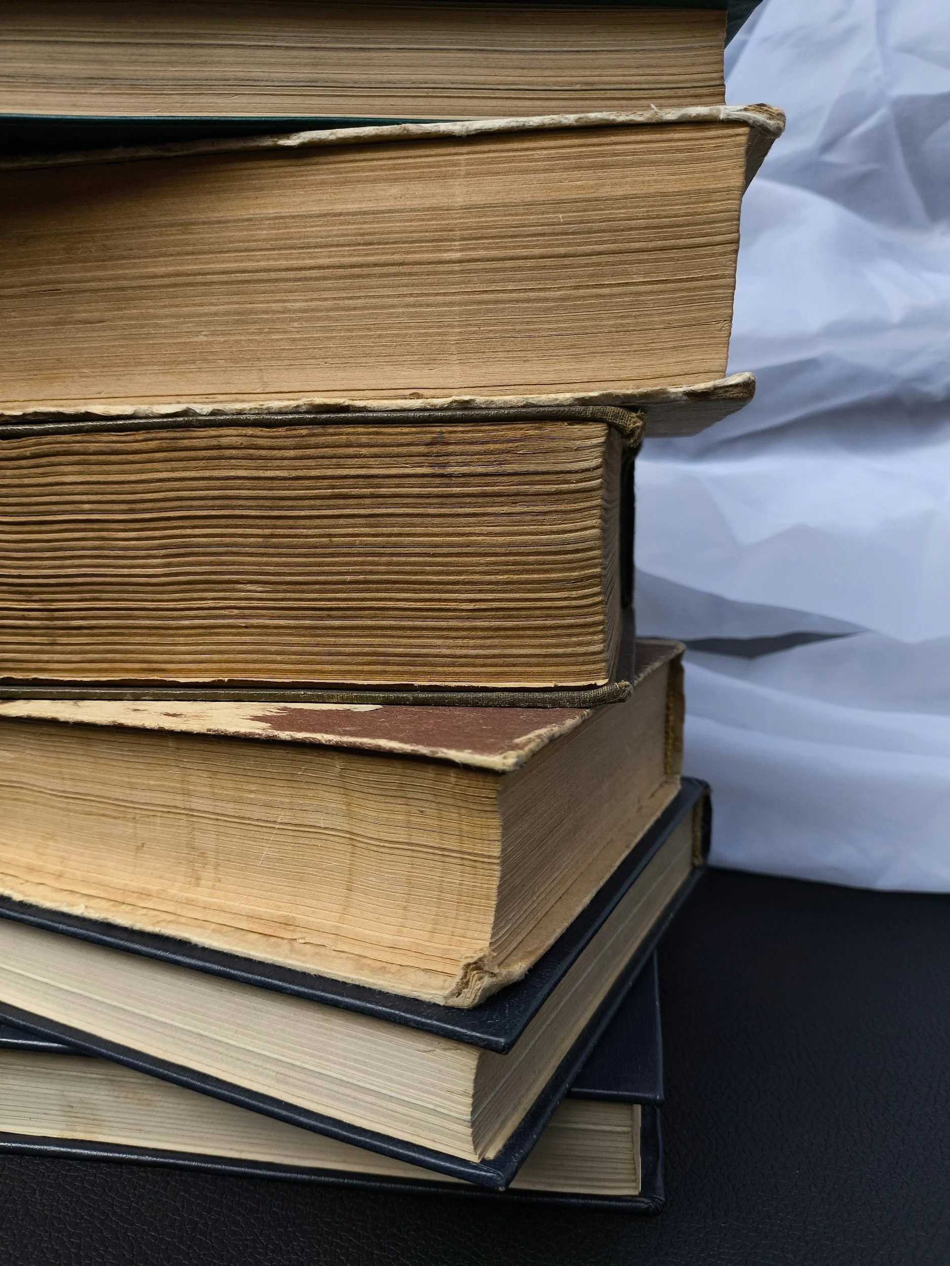 Close-up of a stack of old books with worn and yellowed pages, some with torn covers, placed on a black surface with a crumpled white background.