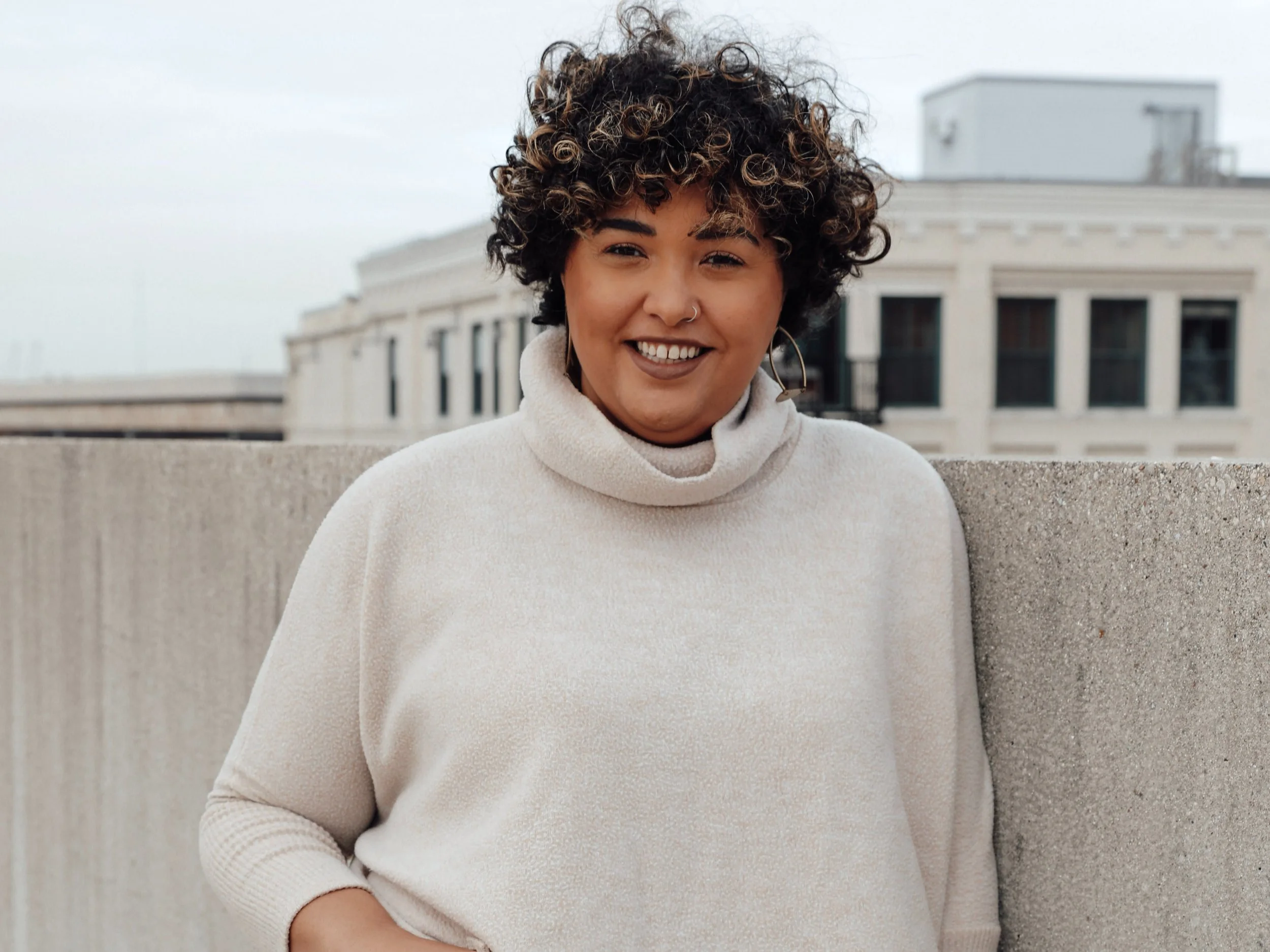 A woman with curly hair smiling outdoors, standing next to a concrete wall with a building in the background.